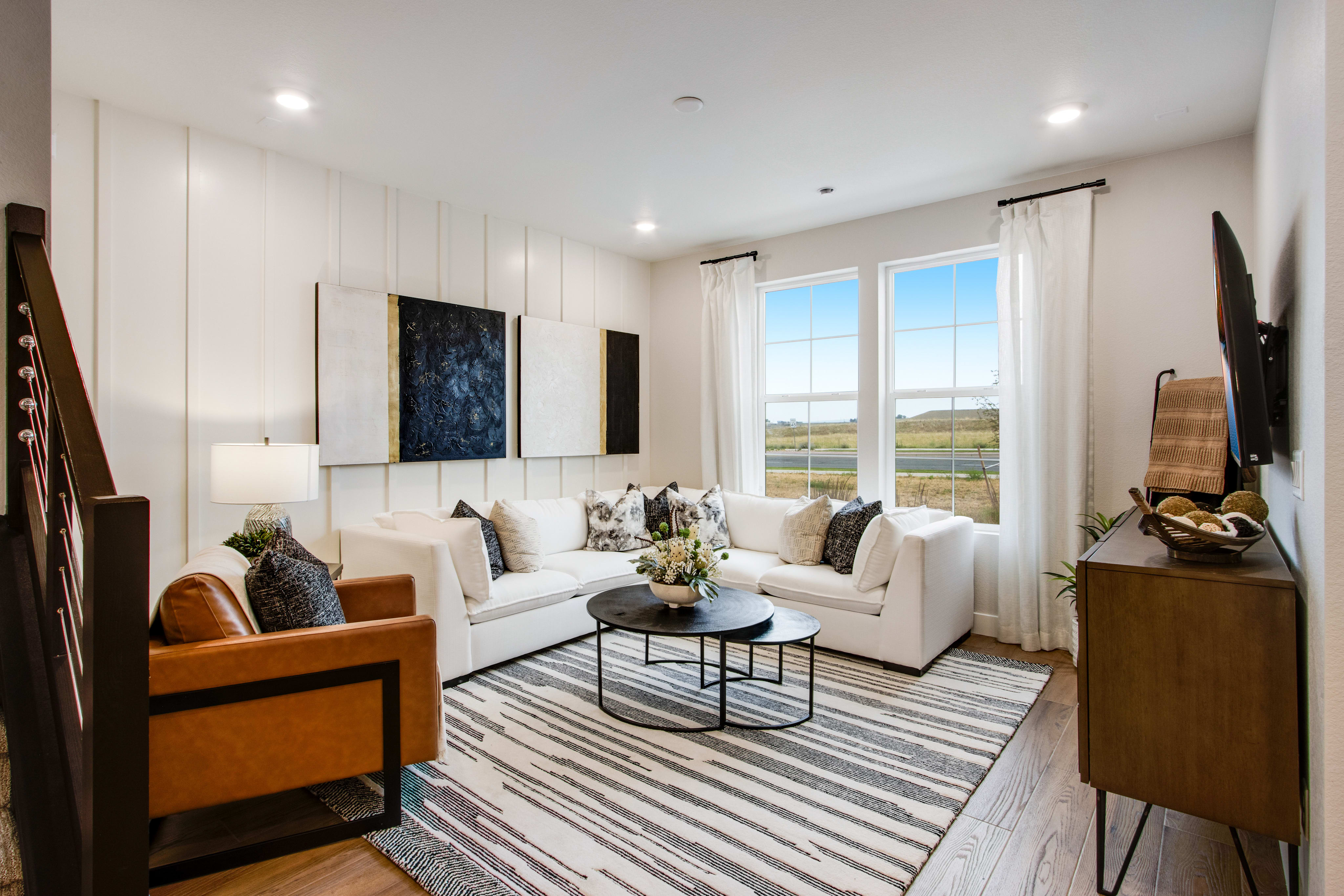 A modern living room with a white sectional sofa and a brown leather armchair in the foreground, and large windows overlooking a scenic landscape in the background.