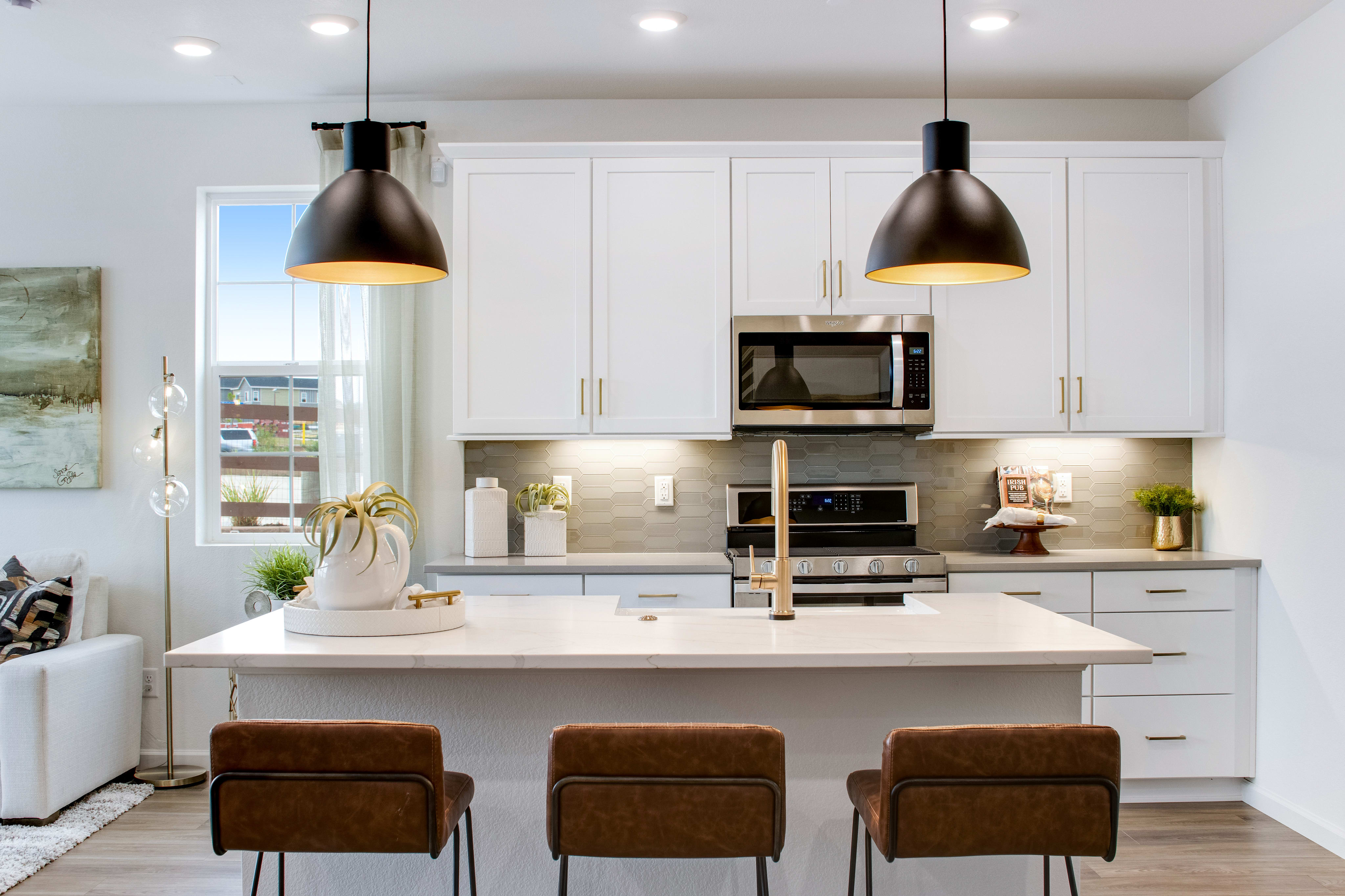 A modern kitchen island with three brown bar stools in the foreground faces white cabinets and stainless steel appliances in the background.