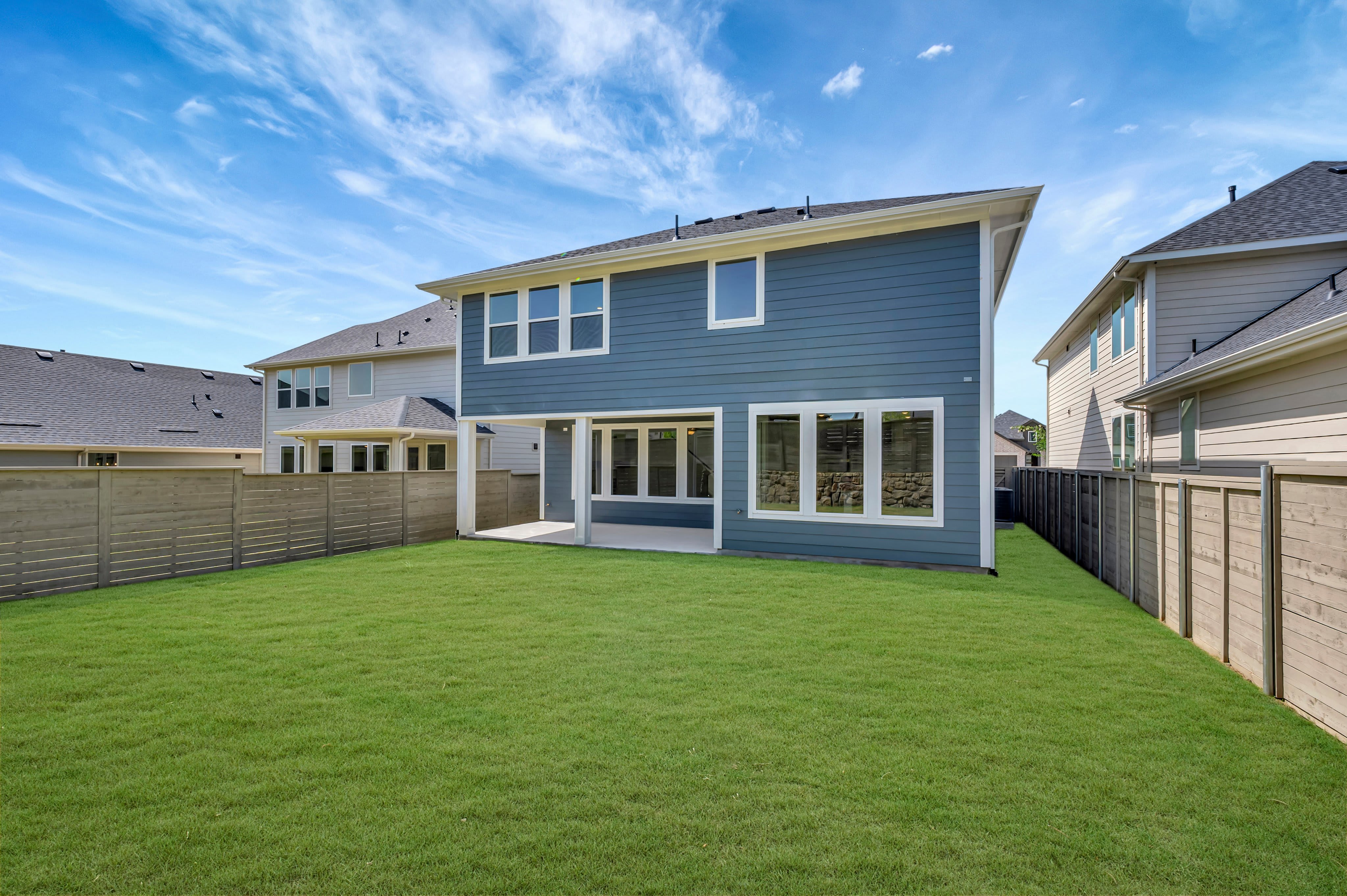 A lush green lawn stretches towards a modern blue house with large windows, while neighboring houses and wooden fences form the background under a bright blue sky.