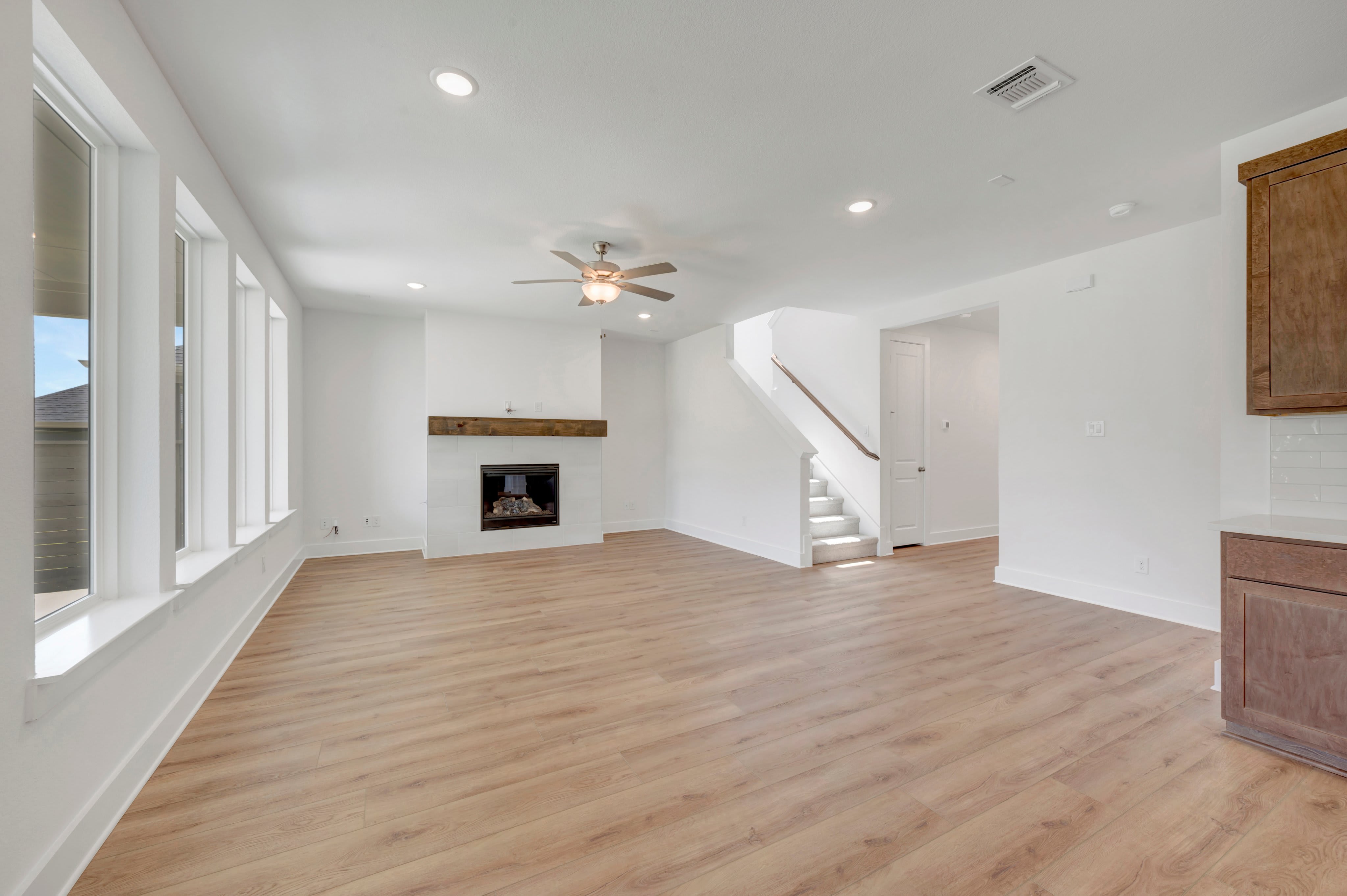 A spacious, modern living room with light wood floors in the foreground and a white fireplace with a wooden mantel in the background.