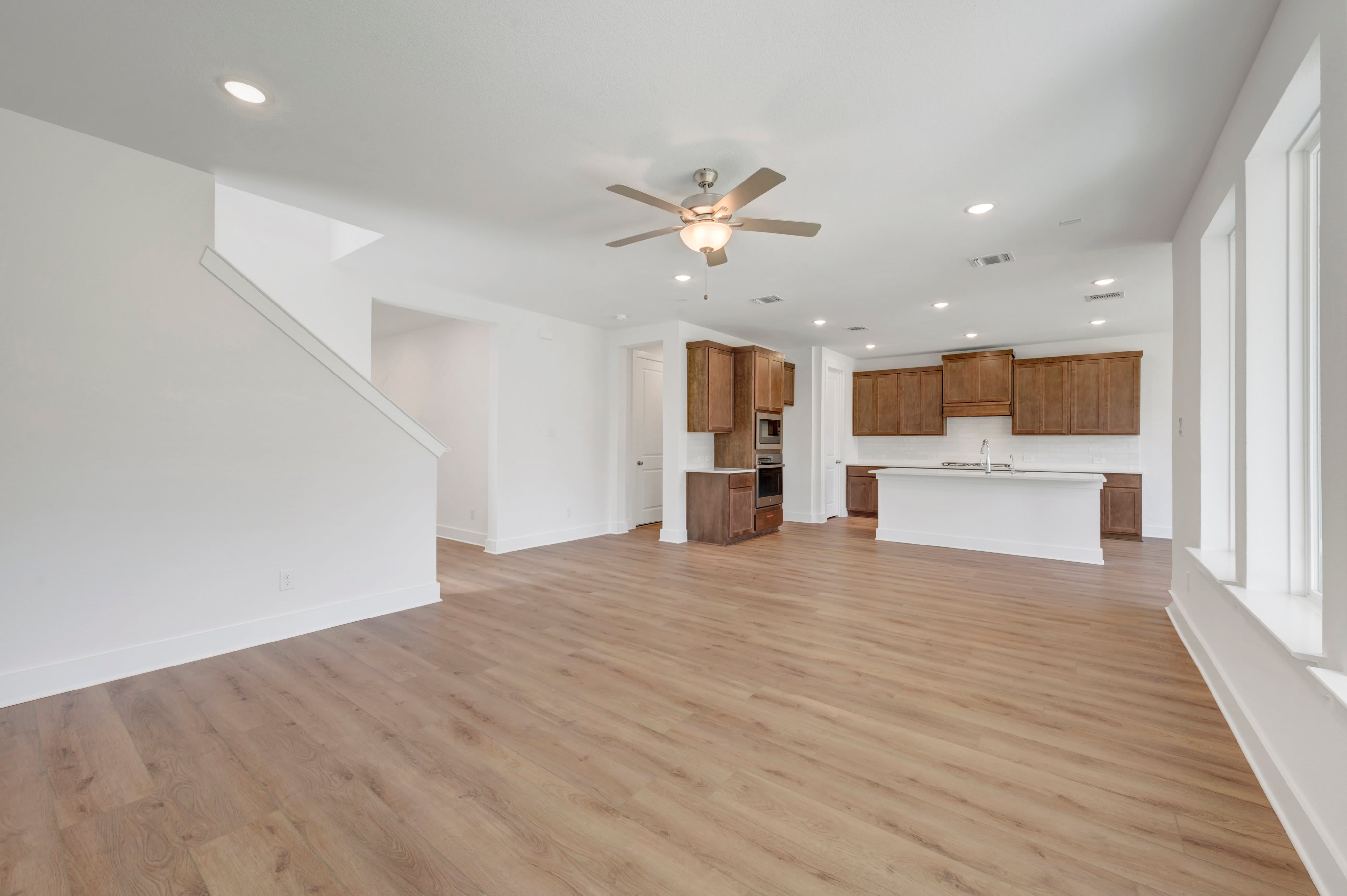 A spacious, modern living area with light wood flooring in the foreground opens to a well-lit kitchen with dark wood cabinets and a white island in the background.