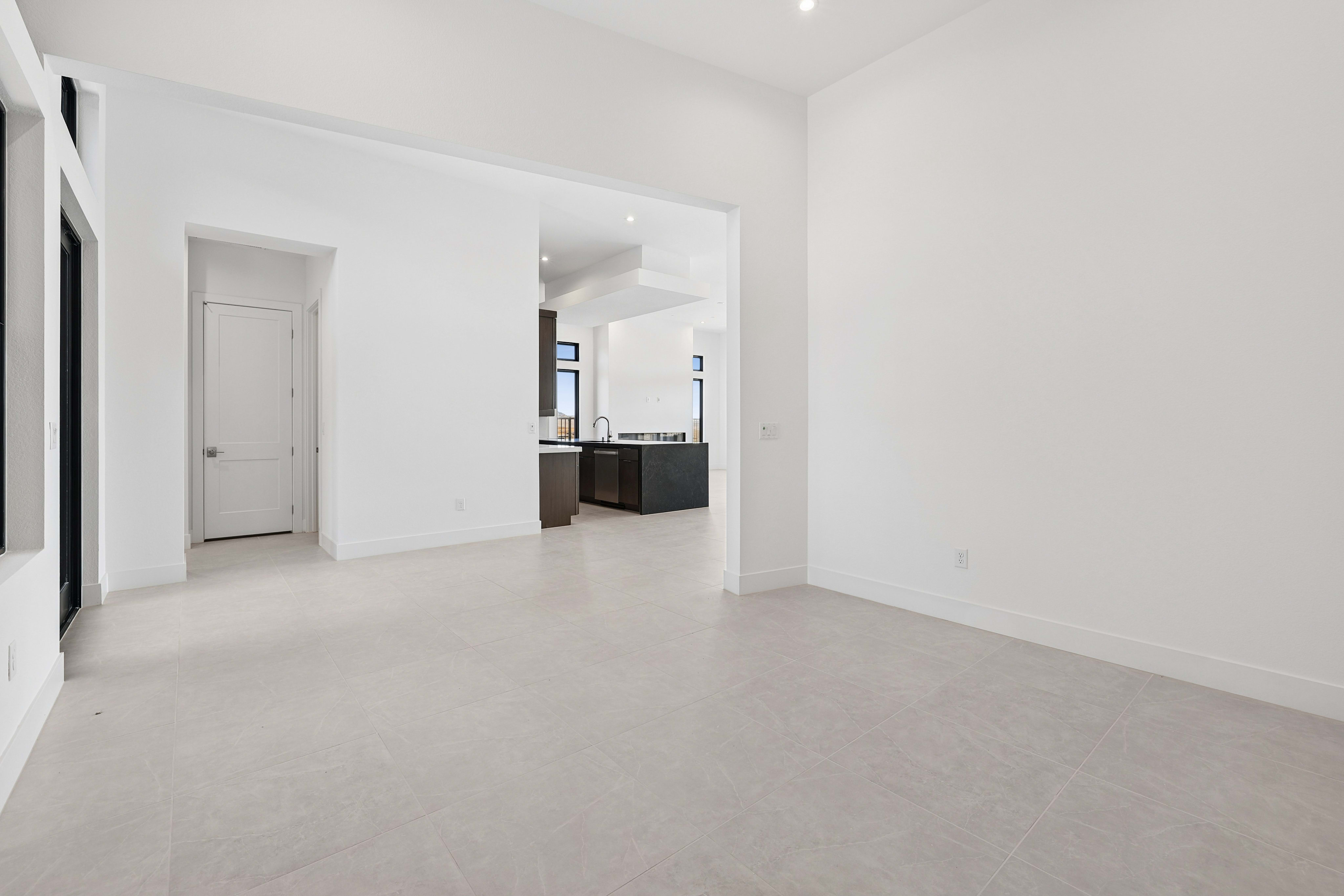 A bright, empty living room with a light carpet in the foreground opens to a modern kitchen with dark cabinetry in the background.