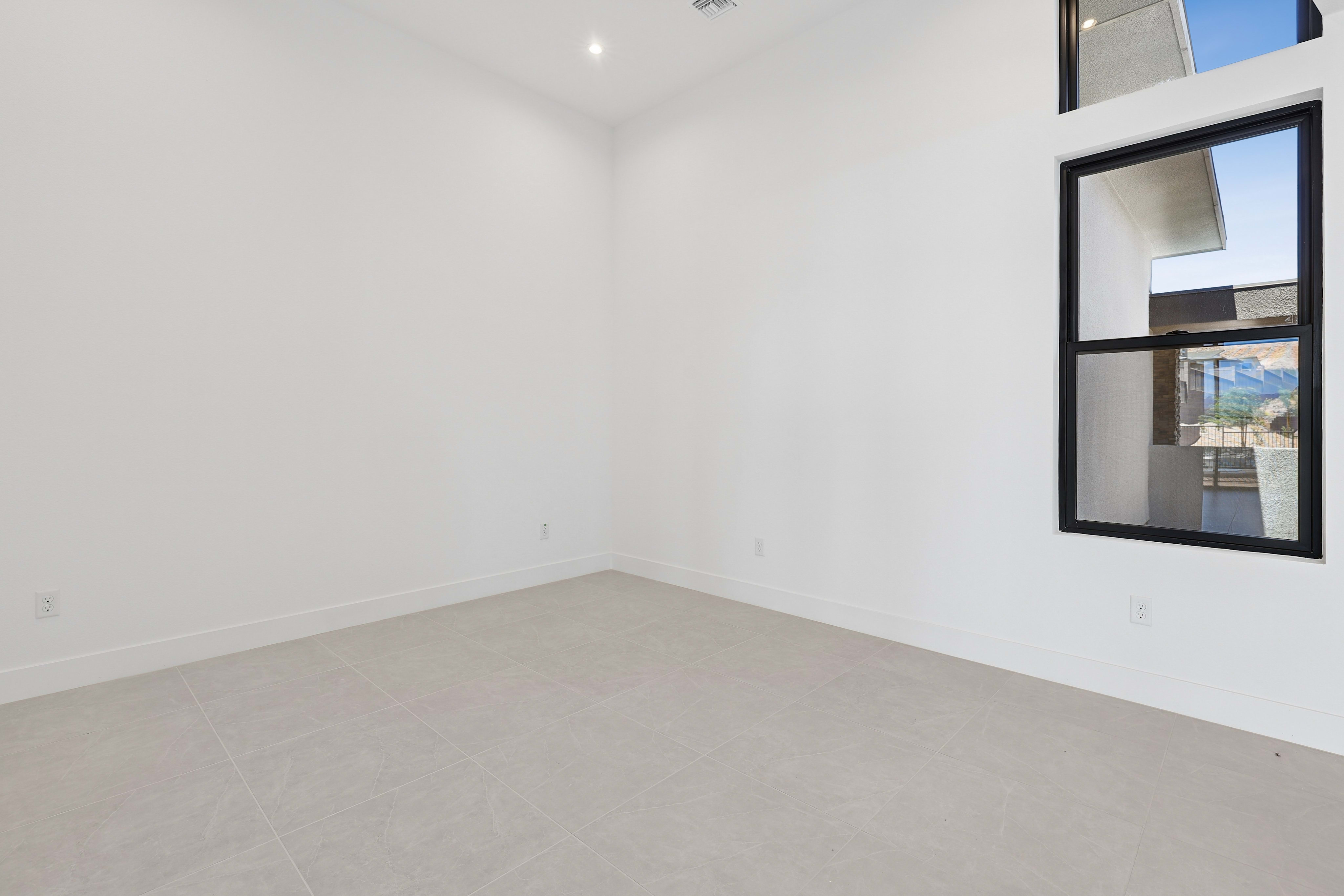 A vacant, modern room with white walls and a beige carpet in the foreground, and a large, multi-paned window with a view of the outdoors in the background.