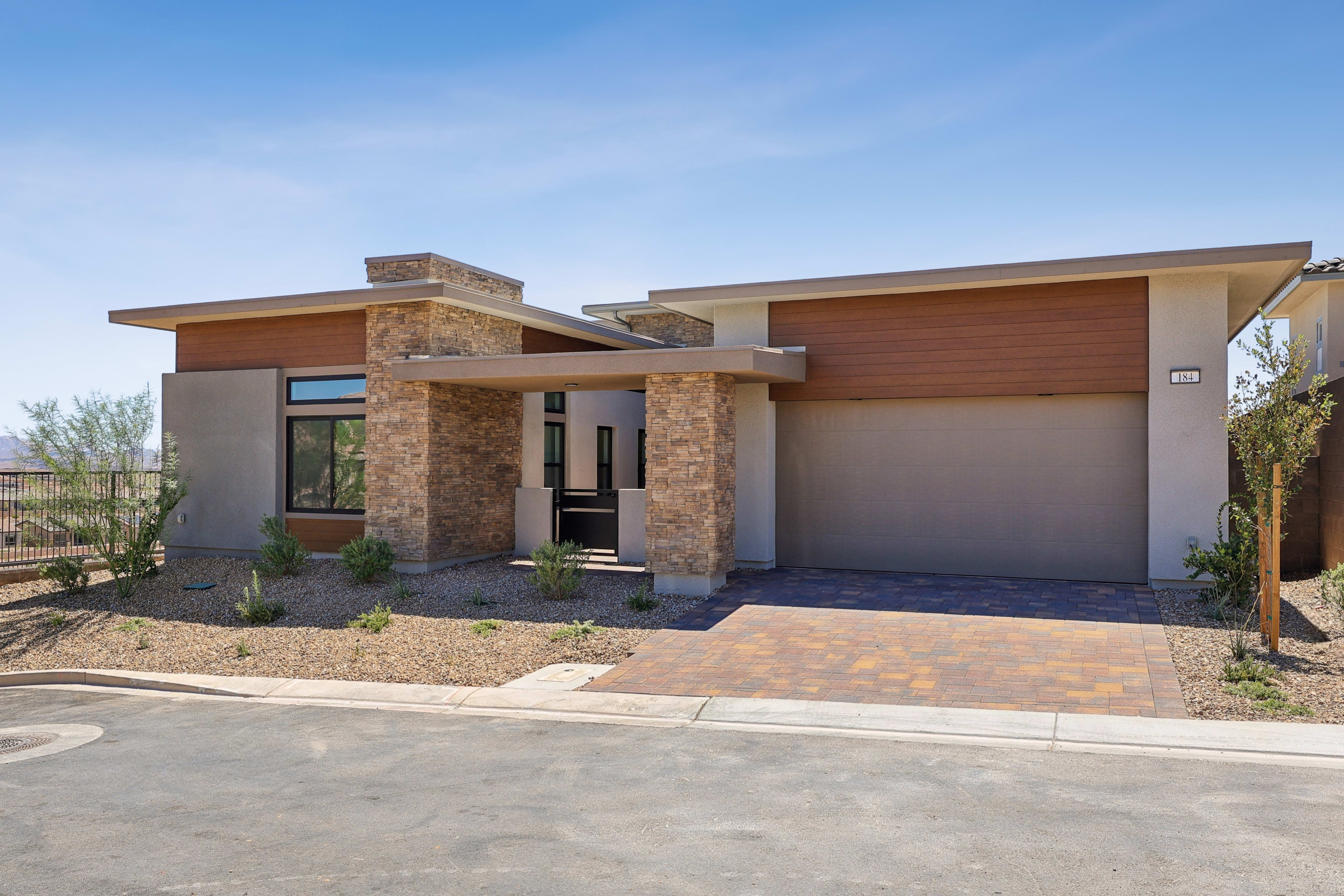 A modern house with stone and wood accents sits on a paved driveway, with a clear blue sky overhead and sparse desert landscaping in the background.