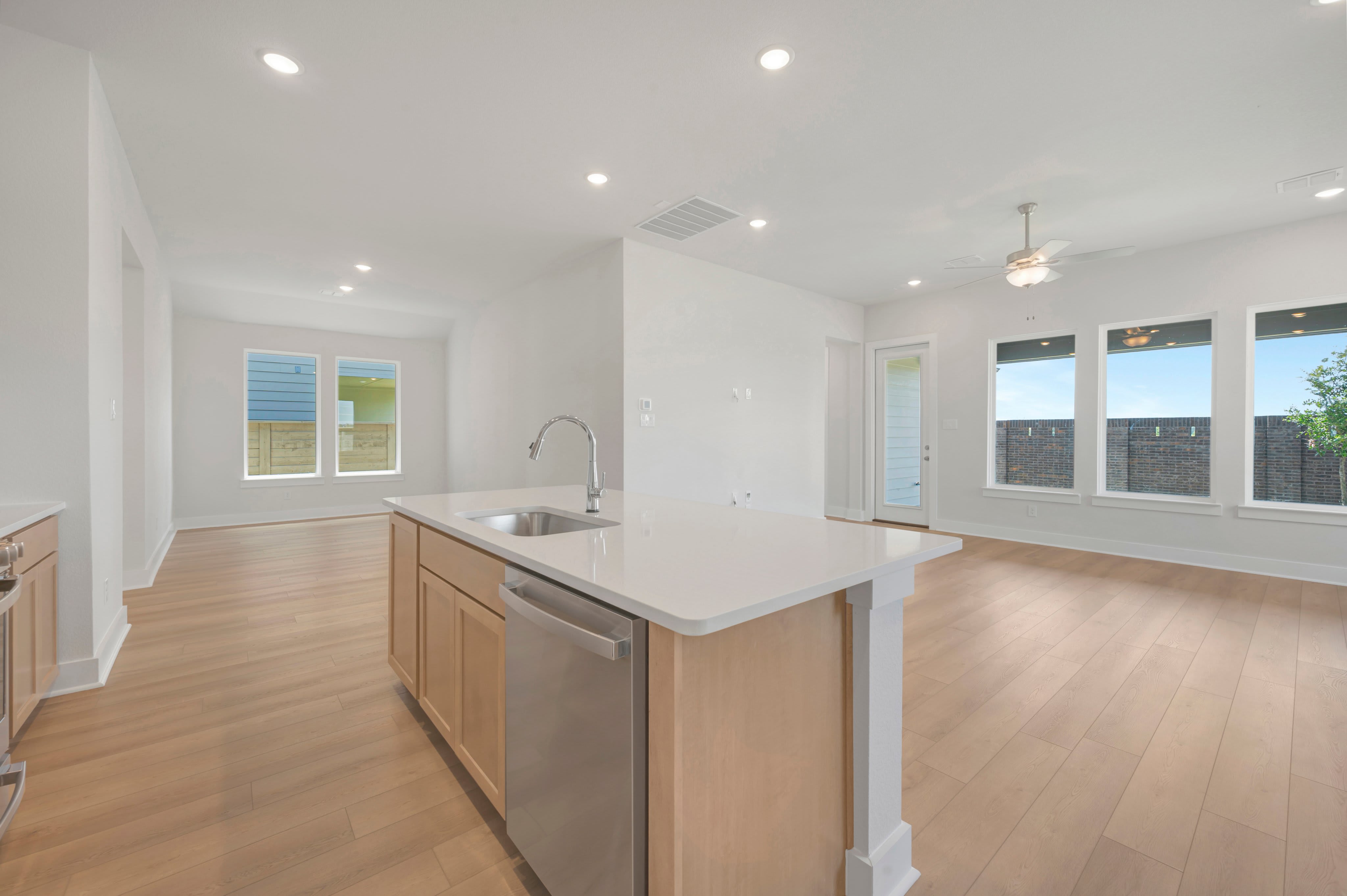 A modern kitchen island with a sink and dishwasher is in the foreground, while a spacious living area with large windows and a ceiling fan is in the background.