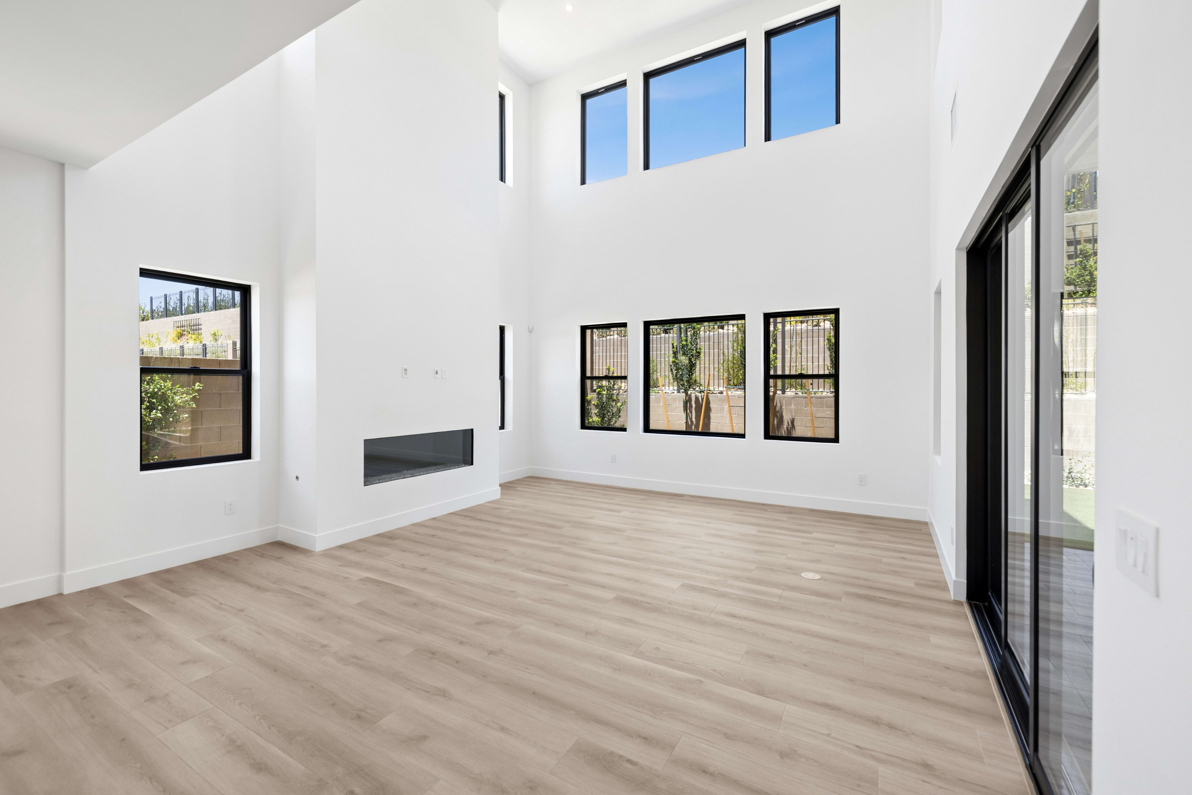 A modern, empty living room with light wood floors in the foreground and large windows revealing a sunny outdoor view in the background.