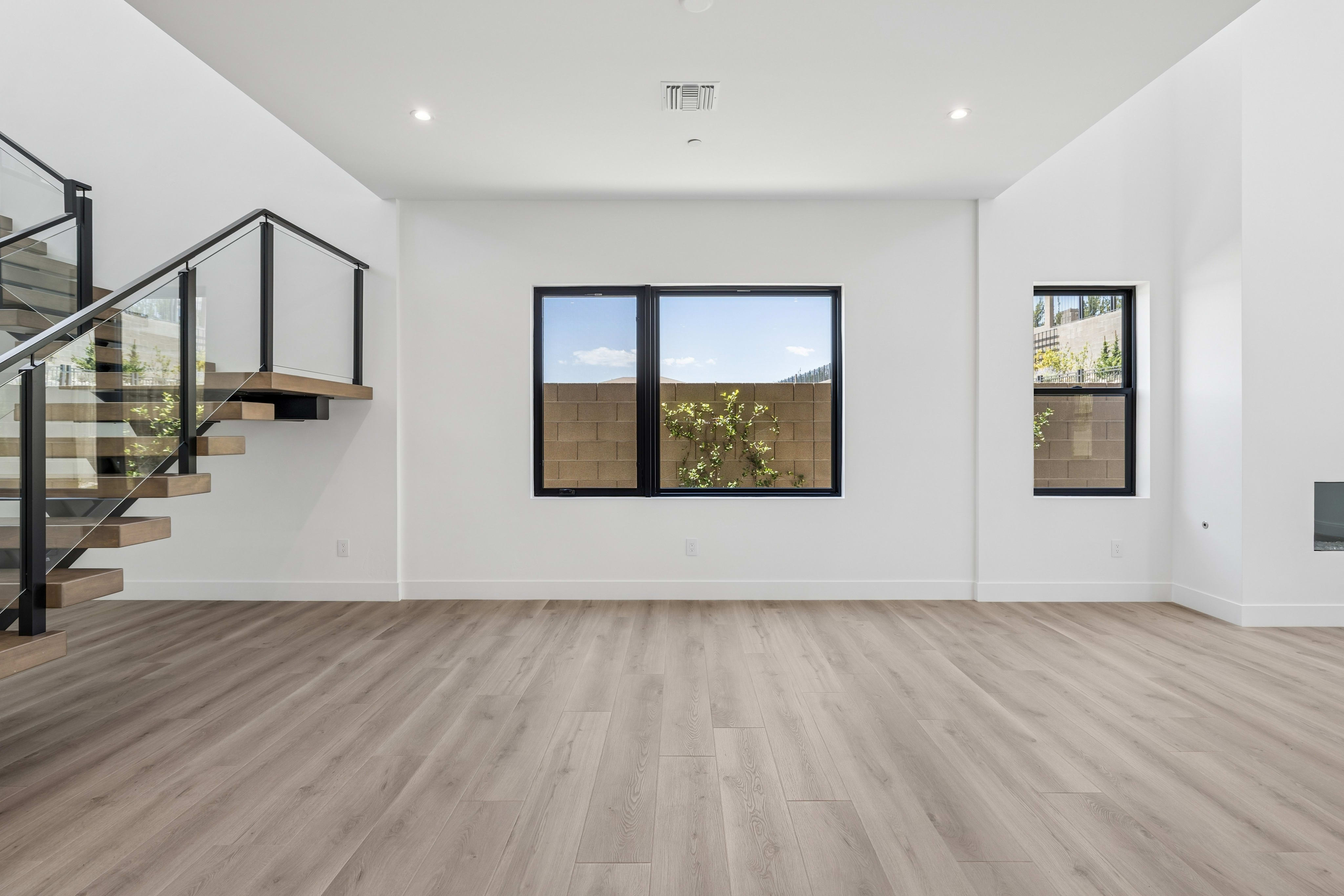 A modern living room with a wooden staircase on the left and large windows revealing a sunny outdoor scene in the background.