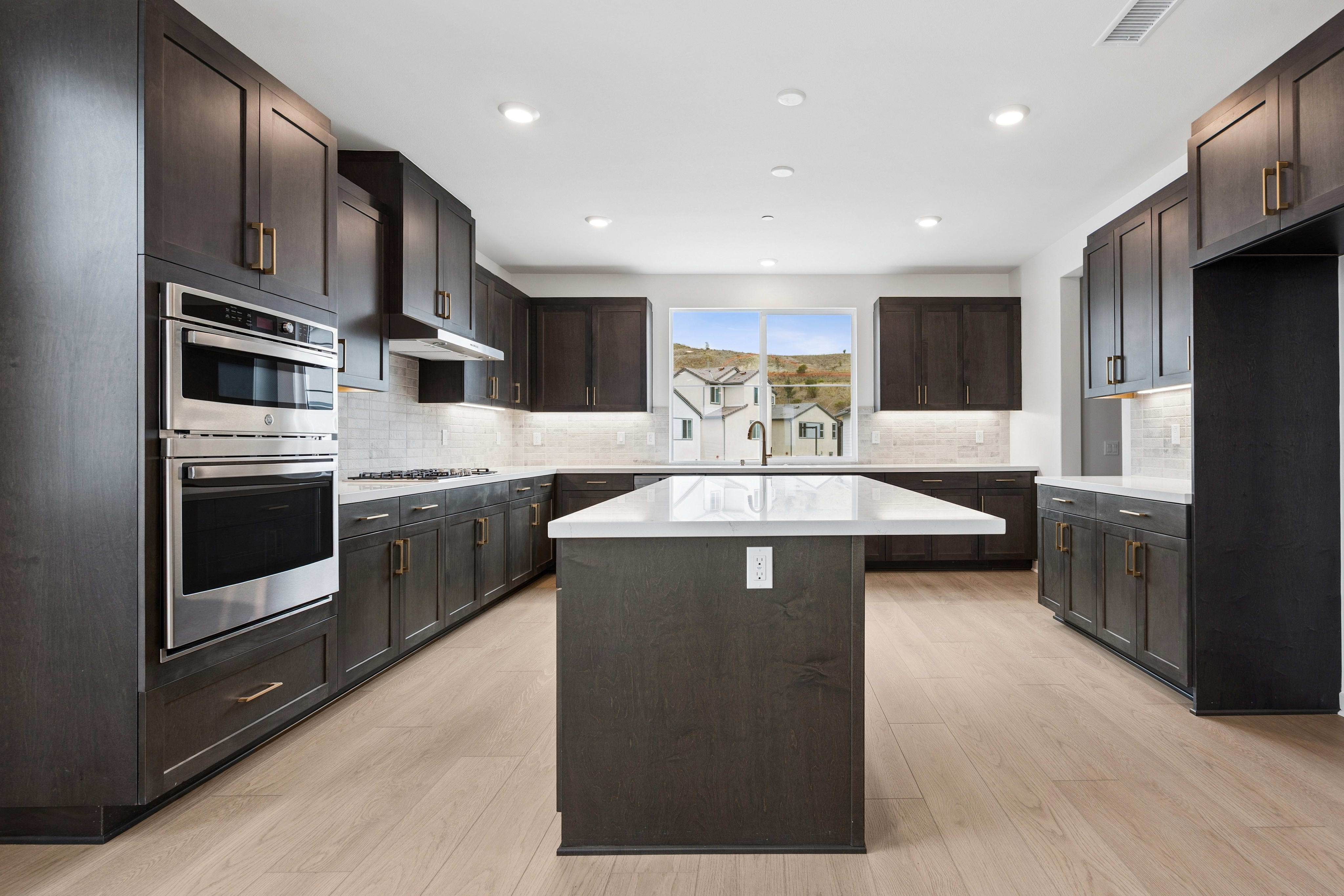 A modern kitchen with dark wood cabinets and a white island in the foreground is set against a background of houses and hills visible through a large window.