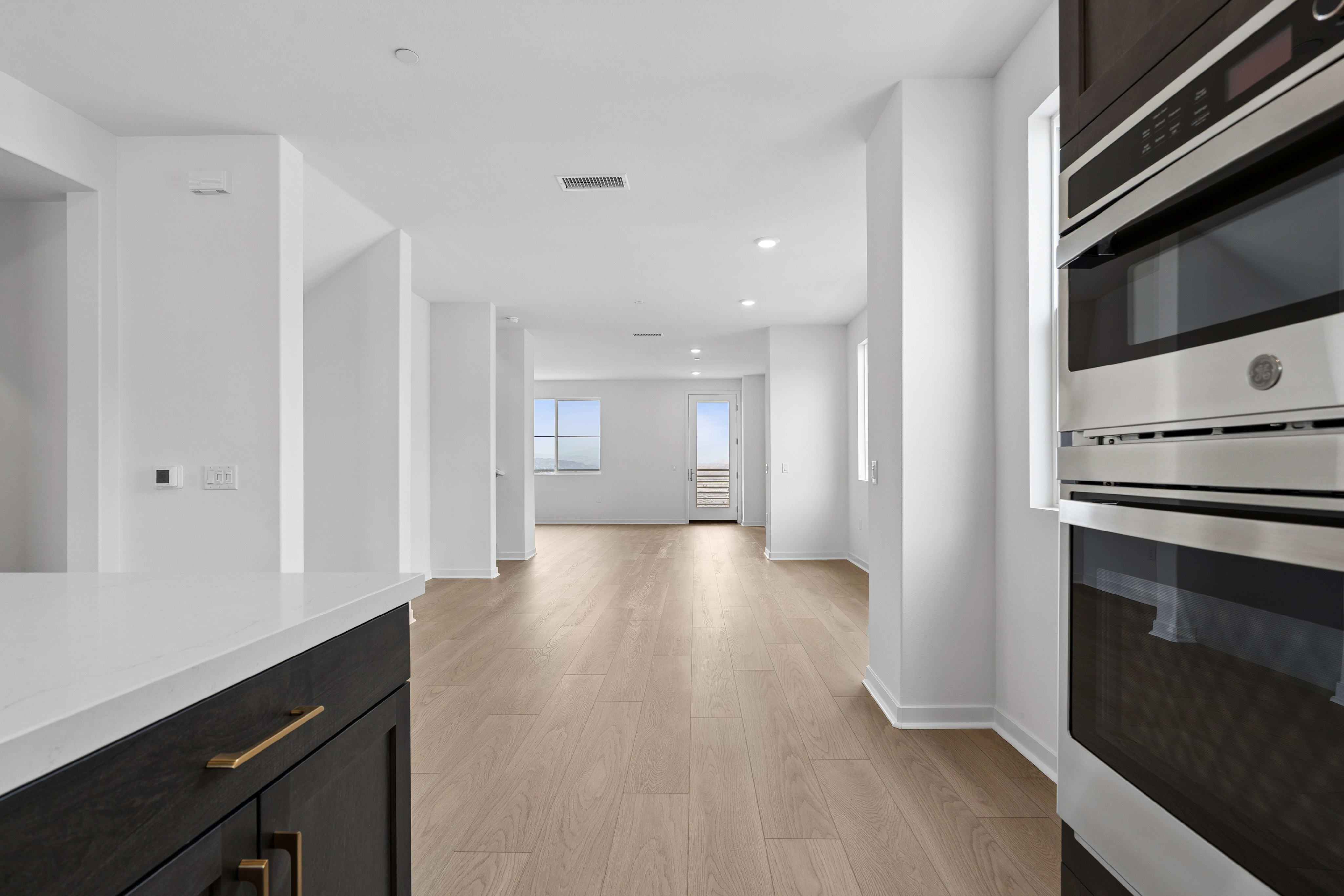 A modern kitchen island with dark cabinets and a white countertop is in the foreground, leading to a bright, open-plan living area with light wood floors and large windows in the background.