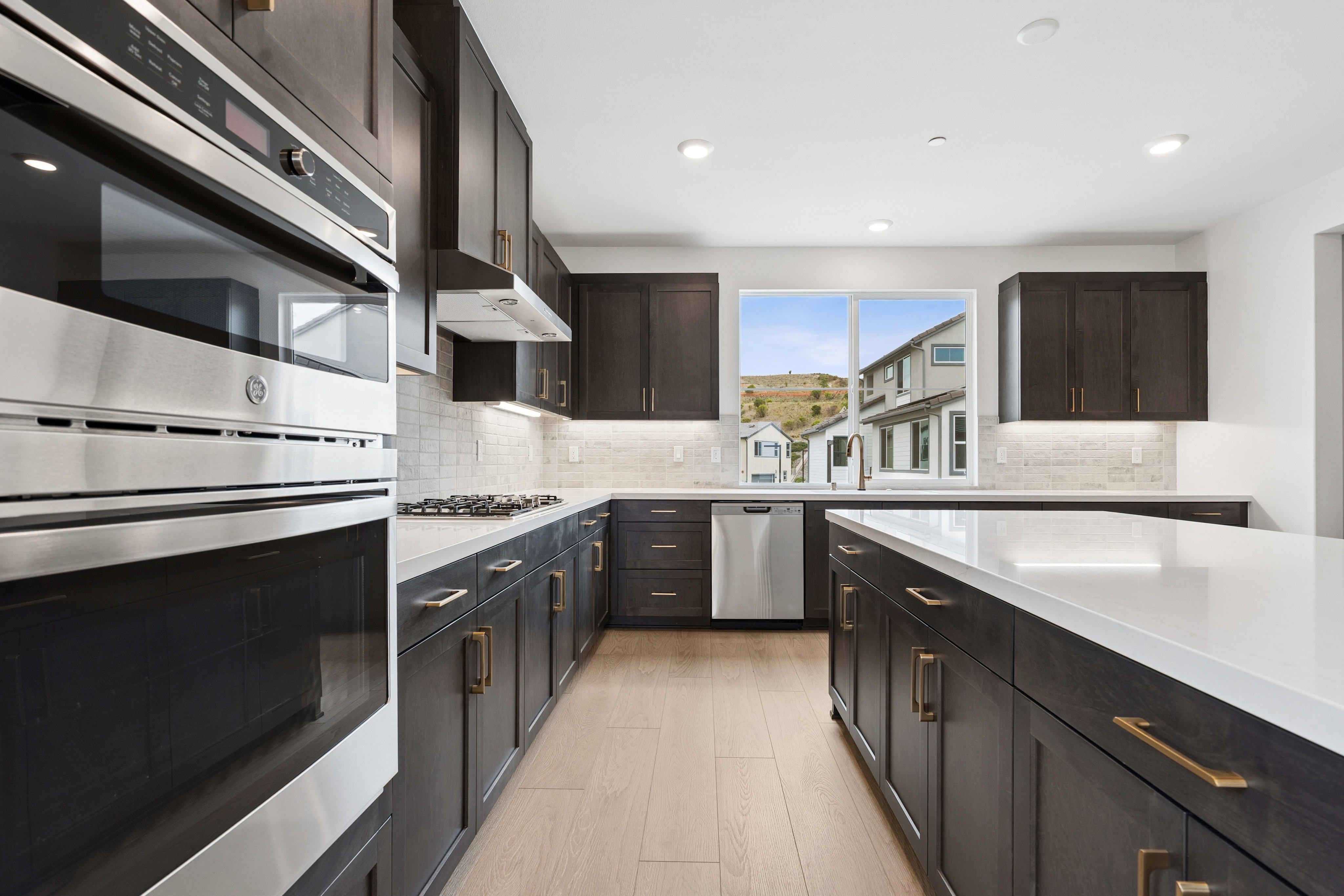 A modern kitchen with dark cabinetry and stainless steel appliances in the foreground is set against a backdrop of a bright window overlooking suburban houses.