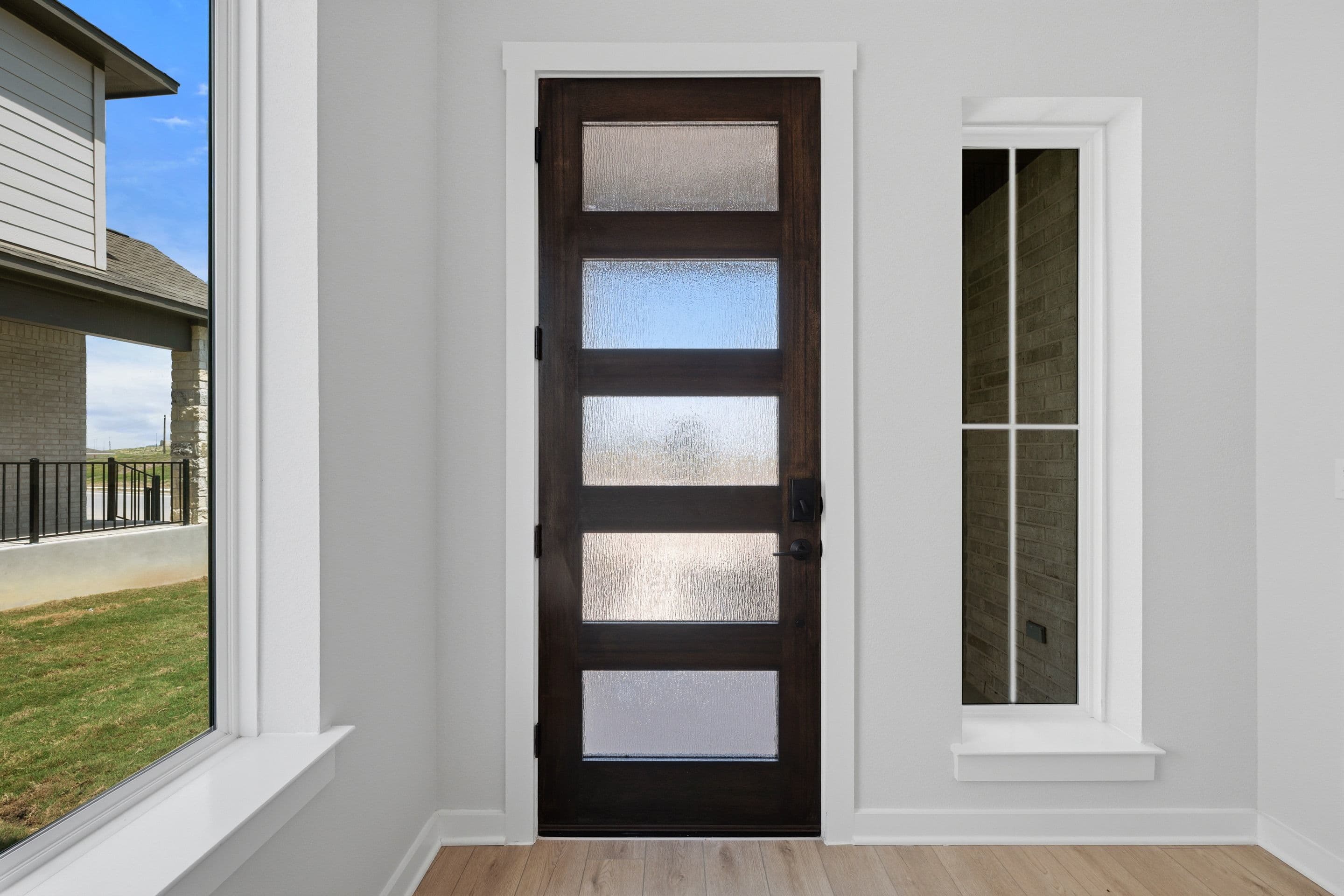 A dark wood door with five frosted glass panels stands in the center, flanked by a tall, narrow window on the right and a wider window on the left, revealing a house and grassy yard in the background.