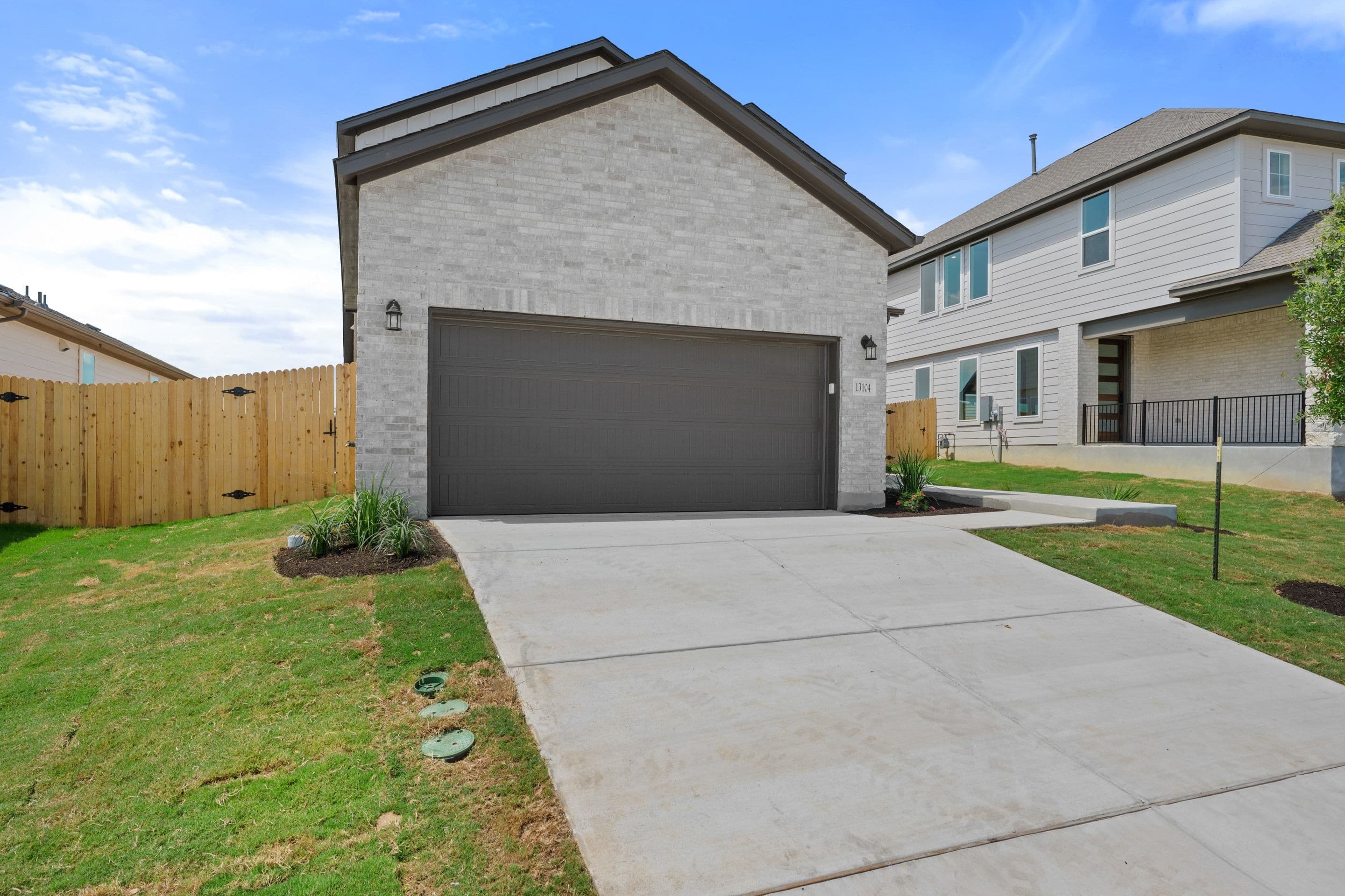 A modern brick house with a gray garage door sits on a concrete driveway, with a wooden fence and green grass in the foreground and other houses in the background.