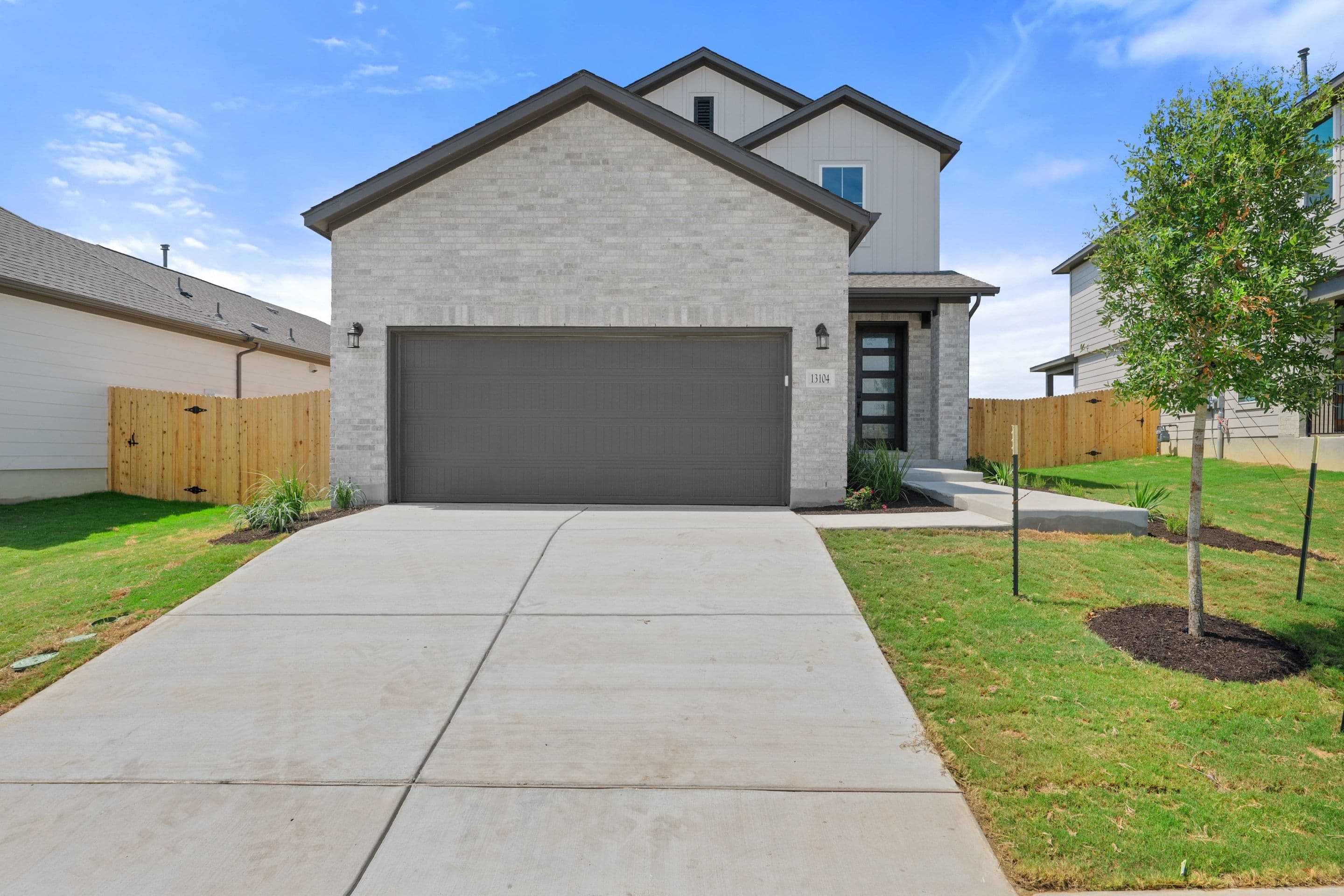 A modern gray garage door dominates the foreground of a light brick house with a black-trimmed entryway, set against a backdrop of a clear blue sky and neighboring houses.