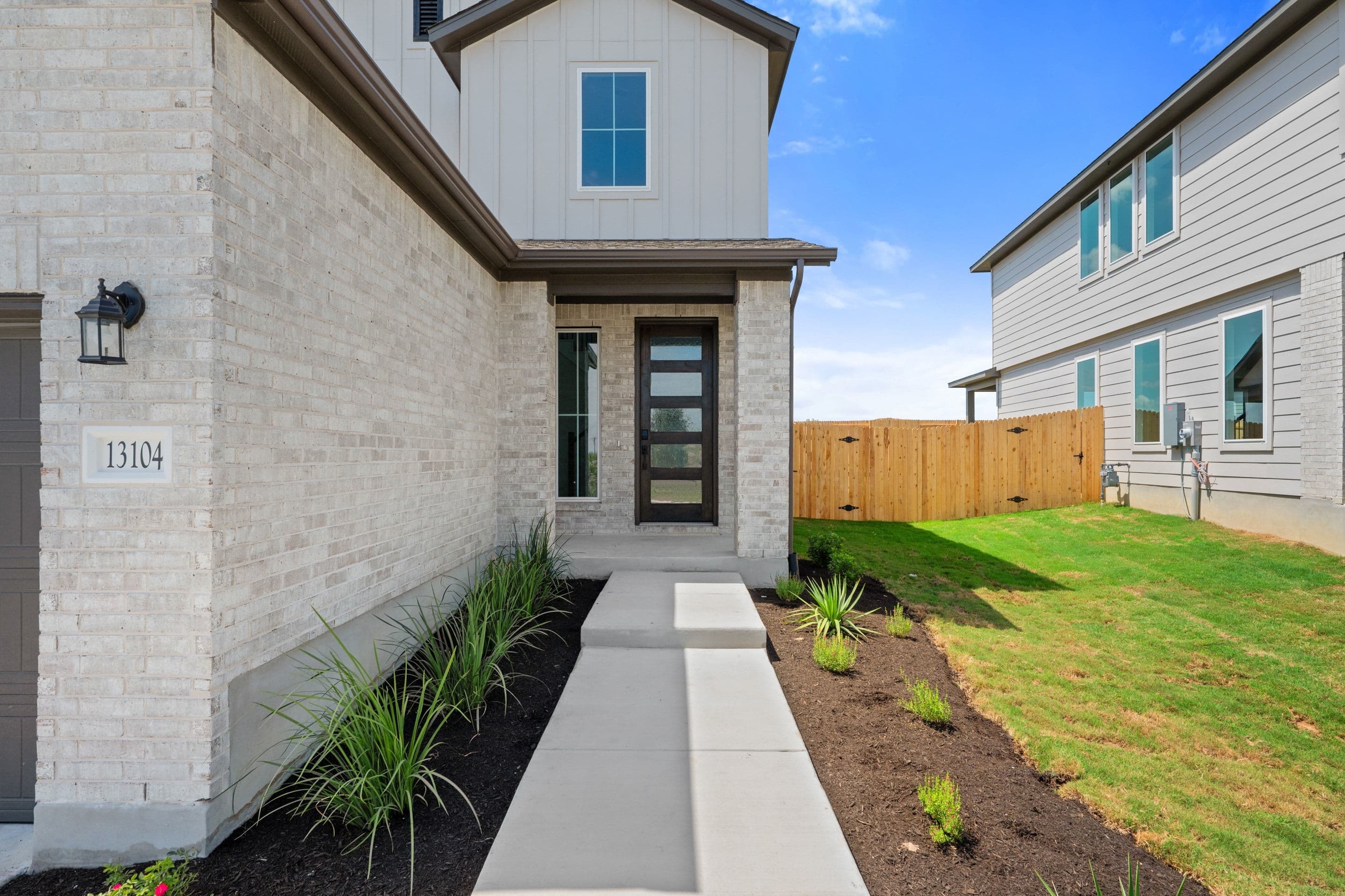 A modern white brick house with a glass-paneled door is approached by a concrete walkway lined with manicured landscaping, while a wooden fence and neighboring houses form the background under a clear blue sky.