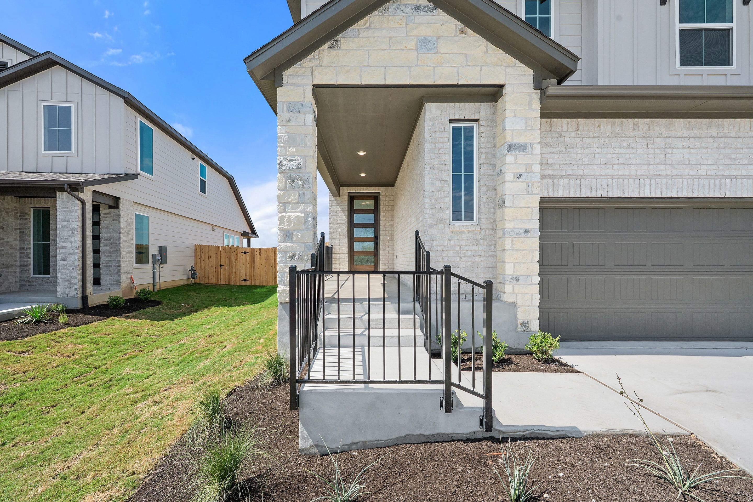 A modern stone-accented house with a black metal railing leading to the front door is situated in a suburban neighborhood with other similar houses and a clear blue sky.