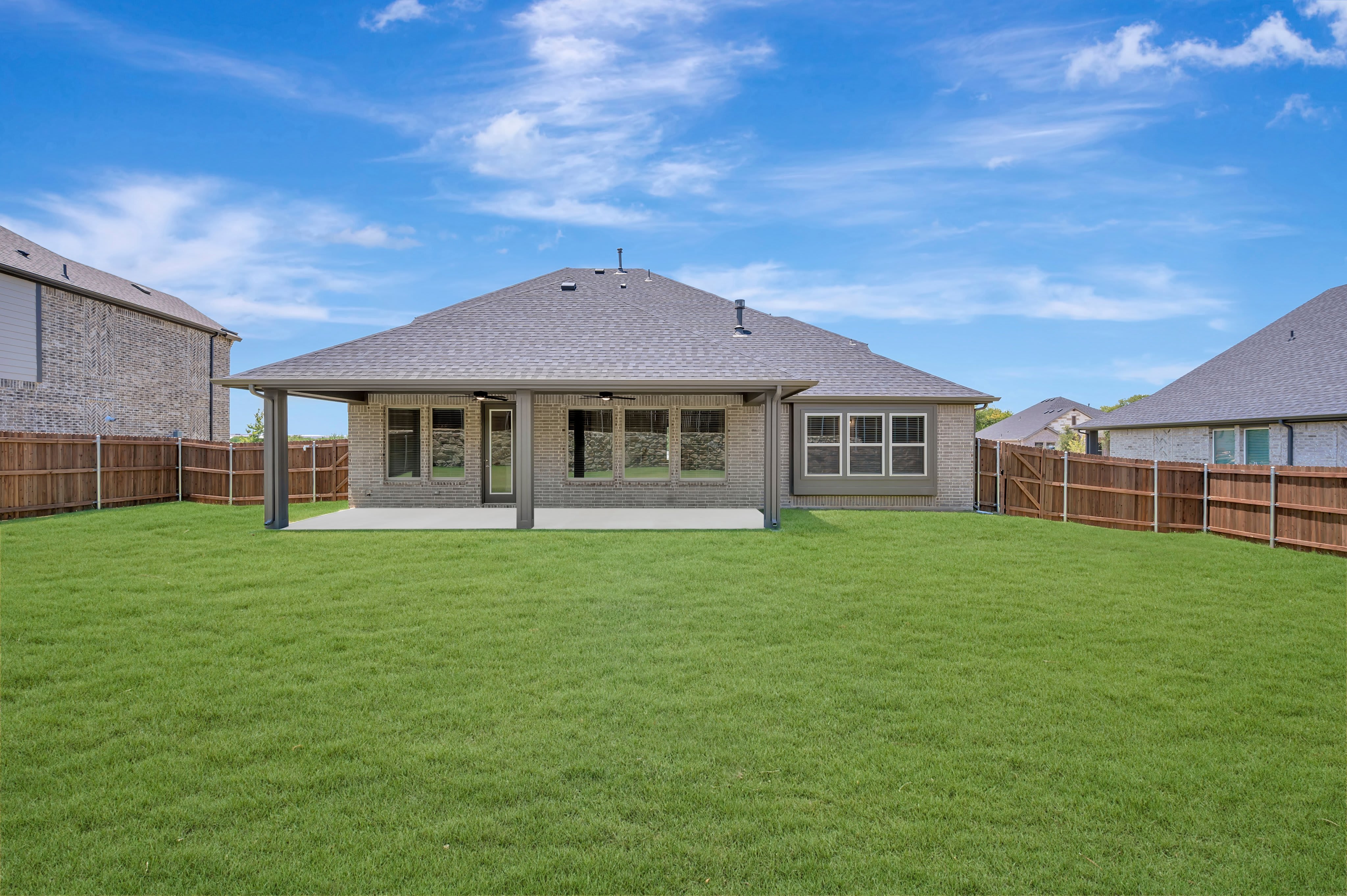 A freshly mowed green lawn stretches out before a modern brick house with a covered patio, all enclosed by a wooden fence under a bright blue sky with scattered clouds.