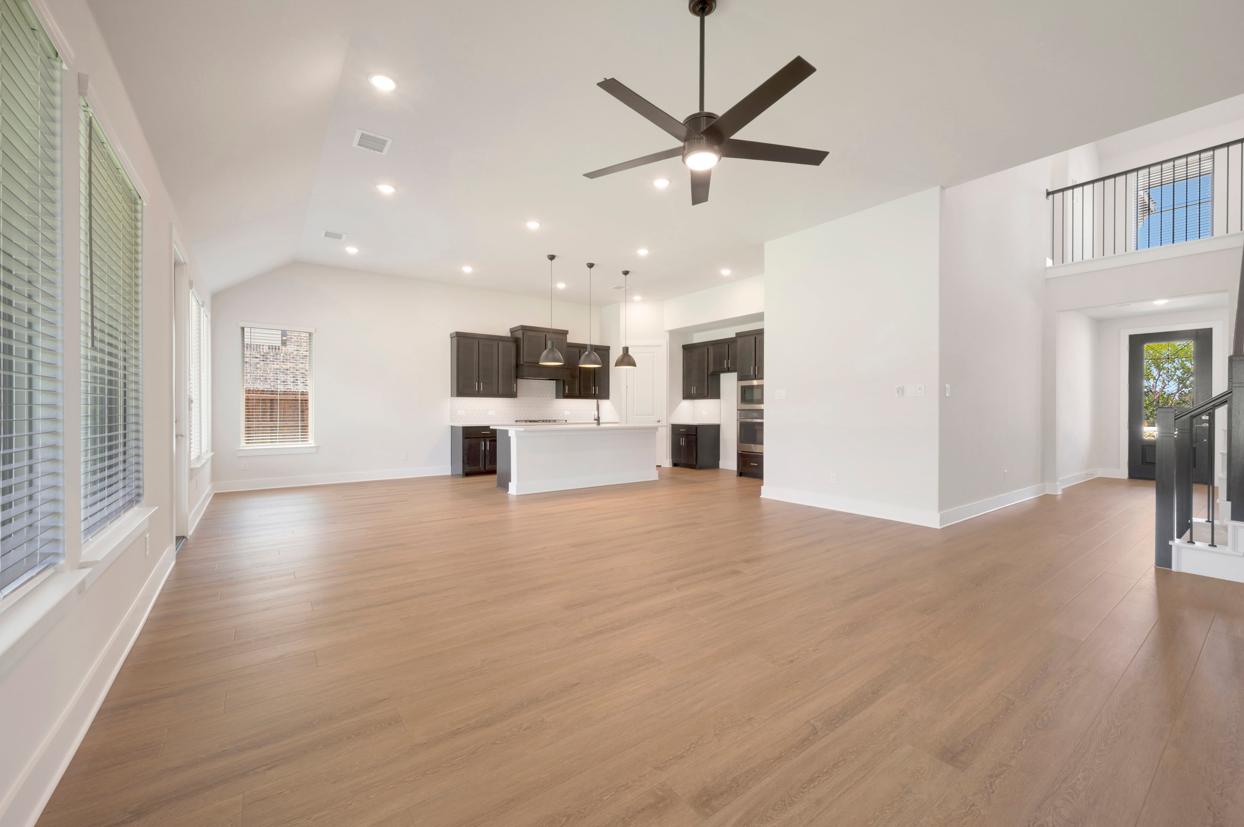 A spacious, modern living area with light wood floors in the foreground opens to a sleek kitchen and a lofted area with a black railing in the background.