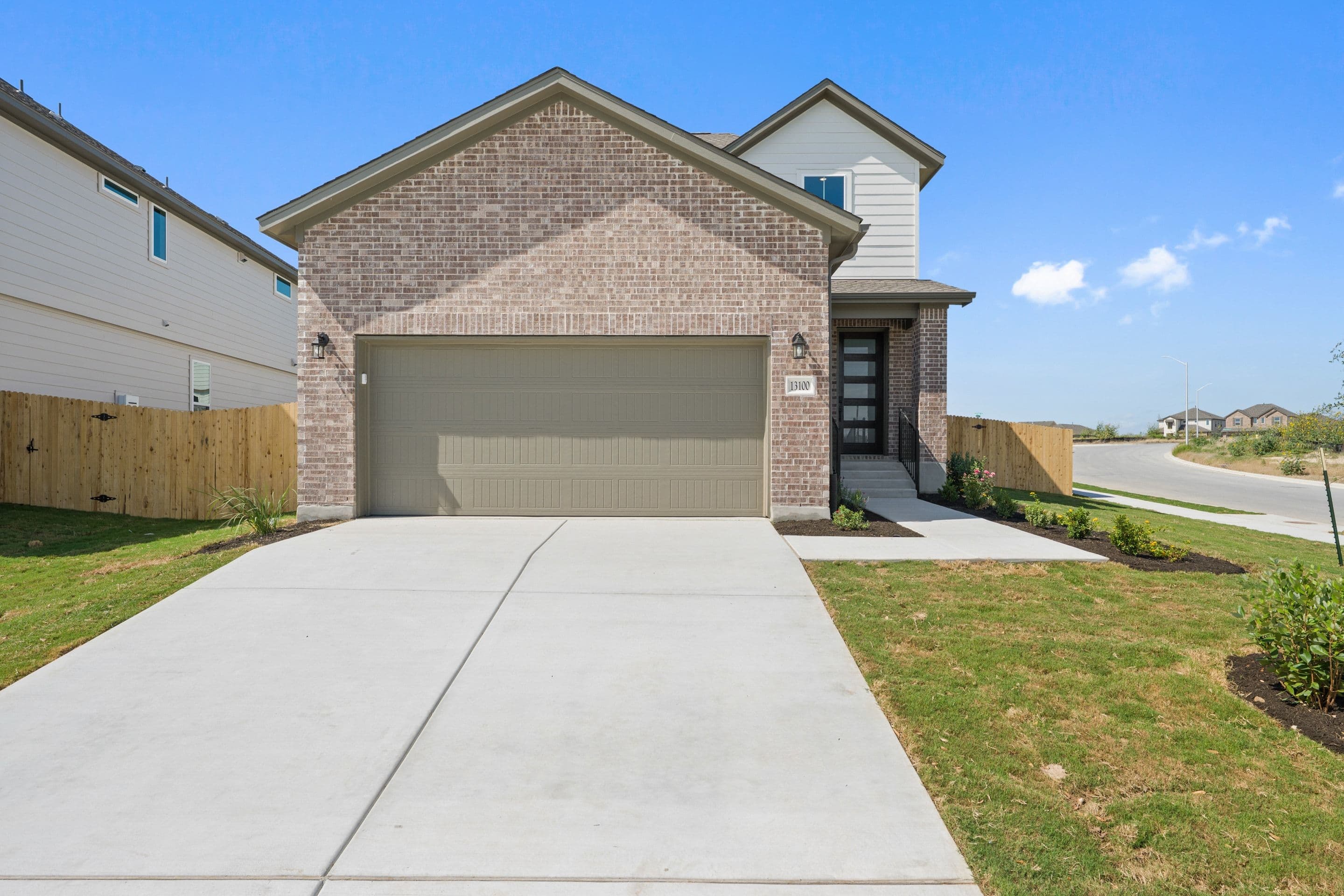 A modern brick house with a gray garage door sits on a concrete driveway in the foreground, while a suburban street curves into the background under a clear blue sky.