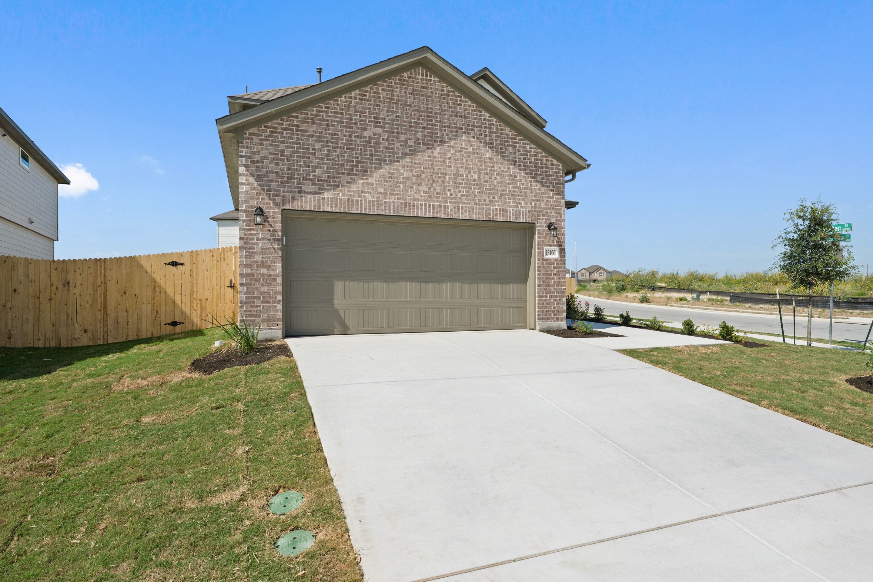 A concrete driveway leads to a brick house with a garage door, bordered by a wooden fence and green grass in the foreground, while a street and distant vegetation are visible in the background under a clear blue sky.