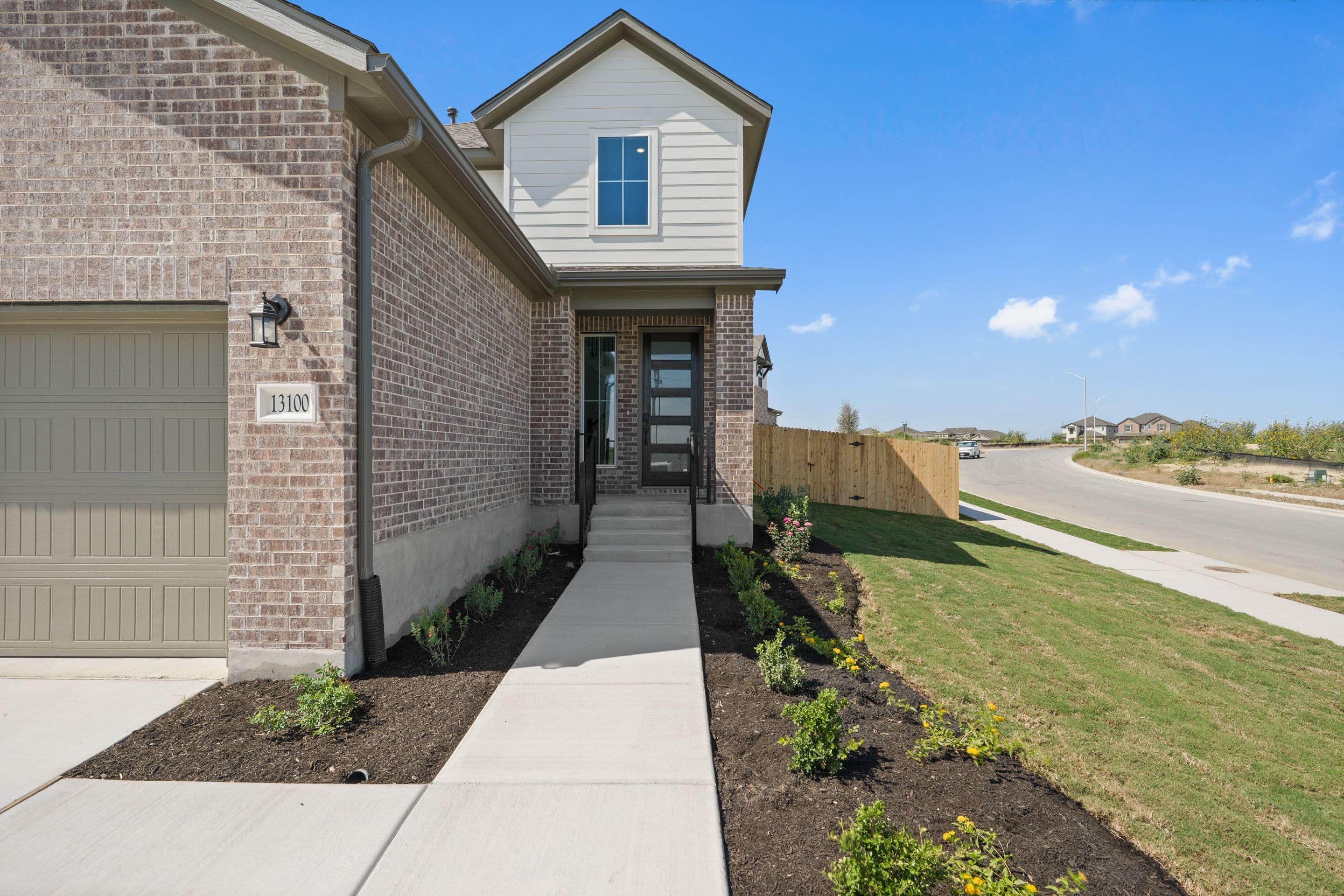 A modern brick house with a paved walkway leading to the front door is surrounded by a manicured lawn and a wooden fence, with a suburban street and more houses visible in the background under a clear blue sky.