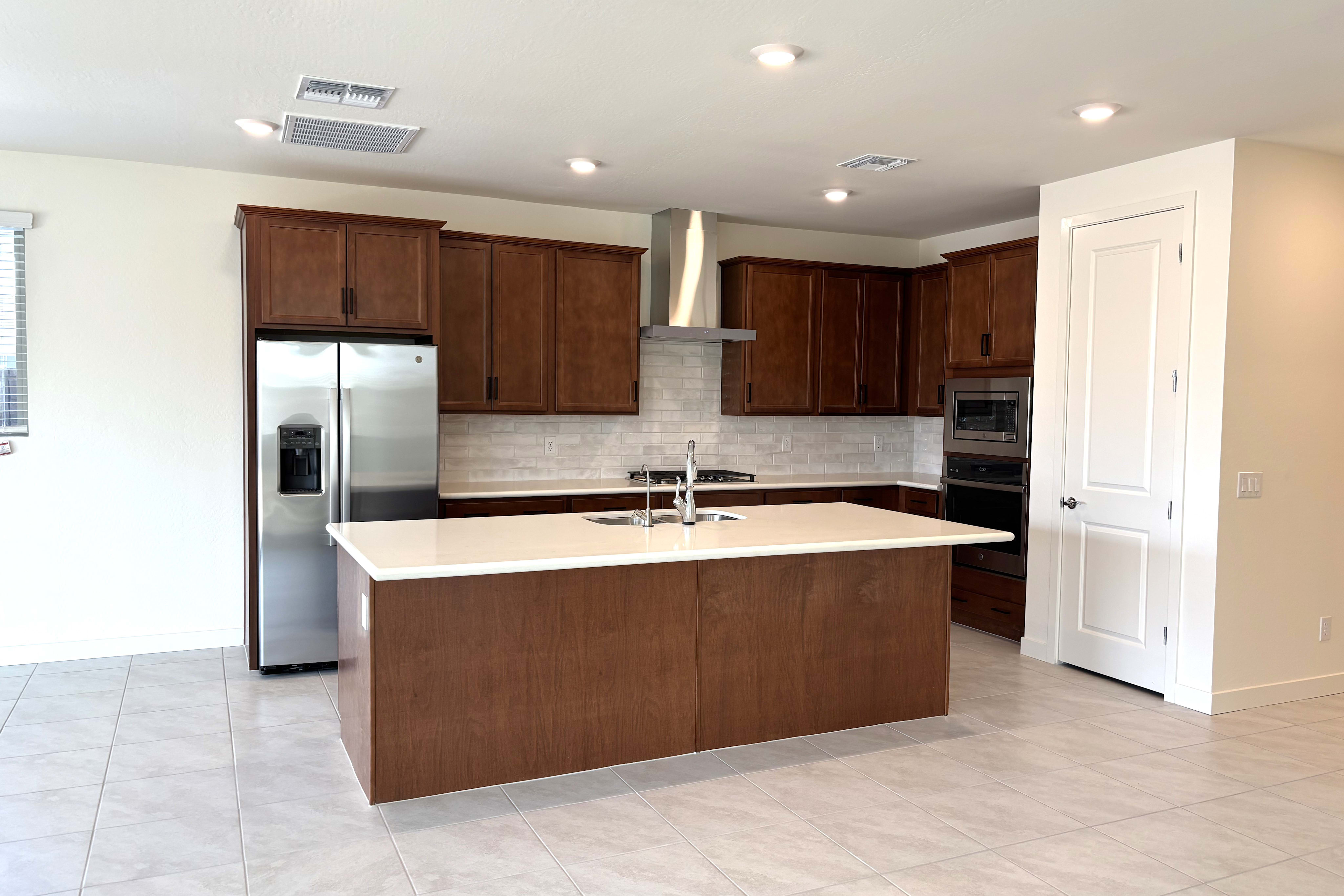 A modern kitchen with a large island in the foreground and dark wood cabinets and stainless steel appliances in the background.