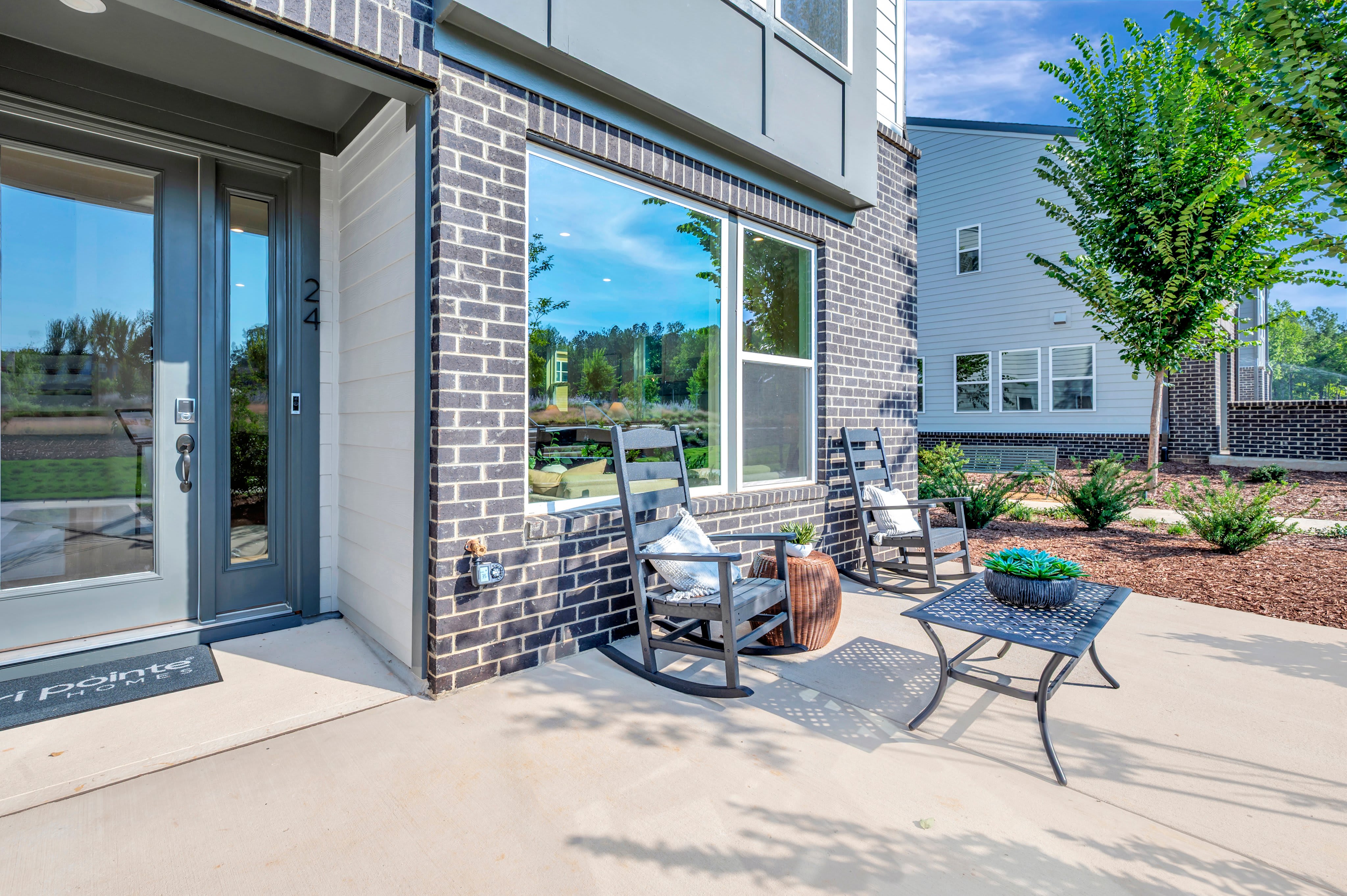 A concrete patio with two rocking chairs and a small table sits in front of a modern brick house, with a lush green landscape and other houses visible in the background.