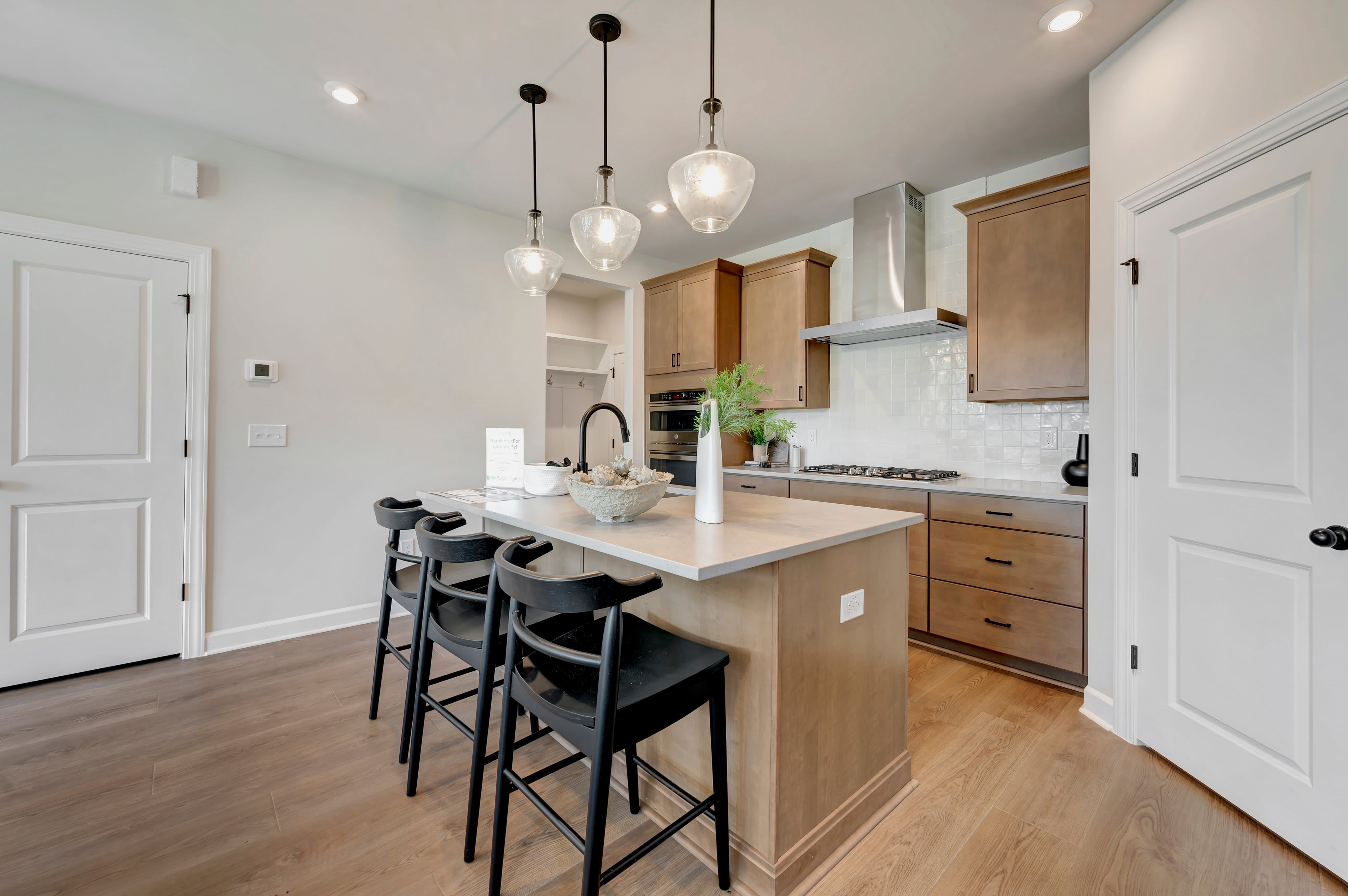 A modern kitchen island with black stools in the foreground is set against a backdrop of wooden cabinets, a tiled backsplash, and stainless steel appliances.