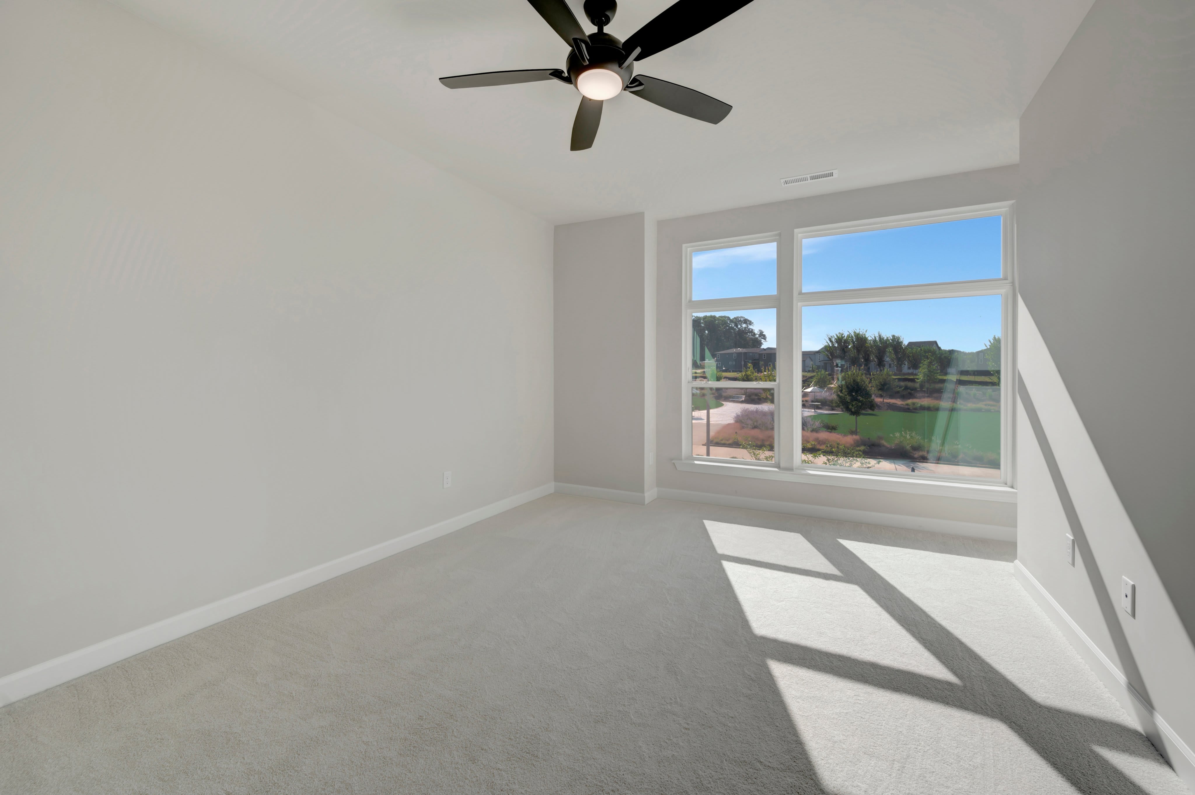 A sparsely furnished room with a ceiling fan in the foreground and a bright window overlooking a green landscape in the background.