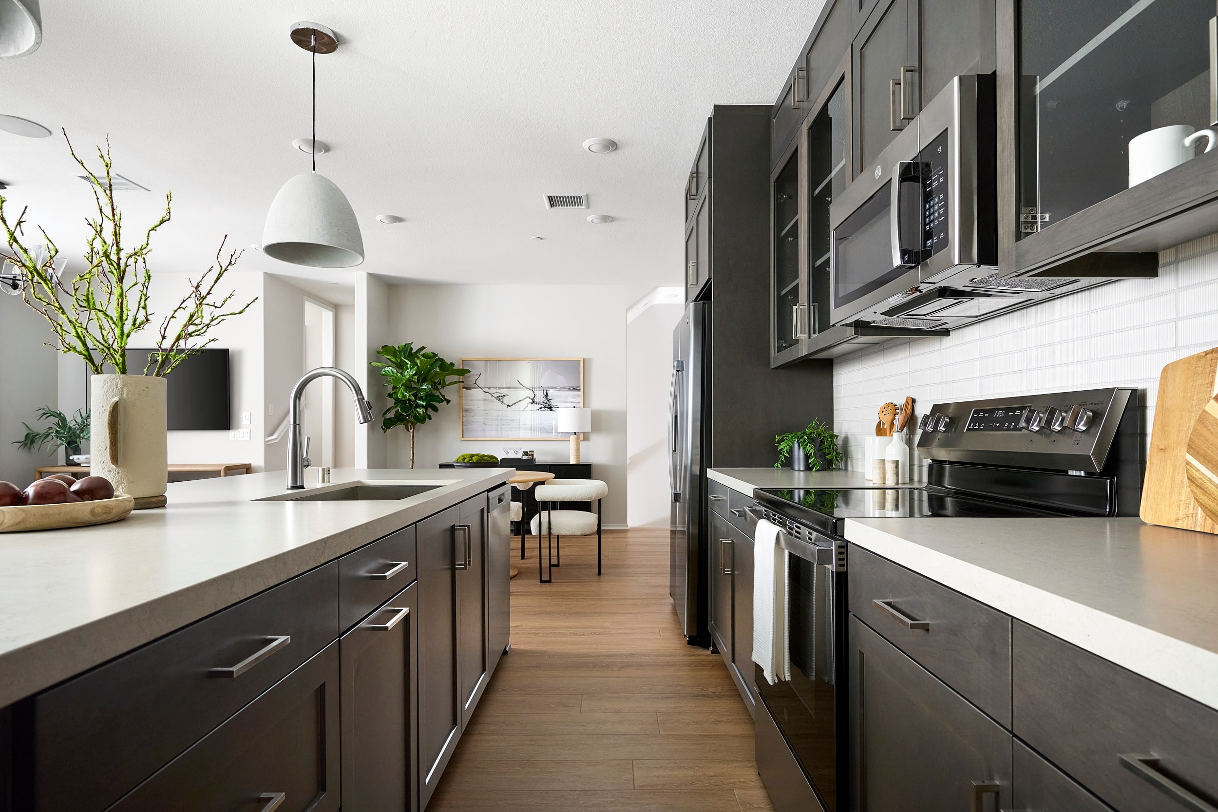 A modern kitchen with dark cabinetry and white countertops in the foreground opens to a dining area with a table and chairs in the background.