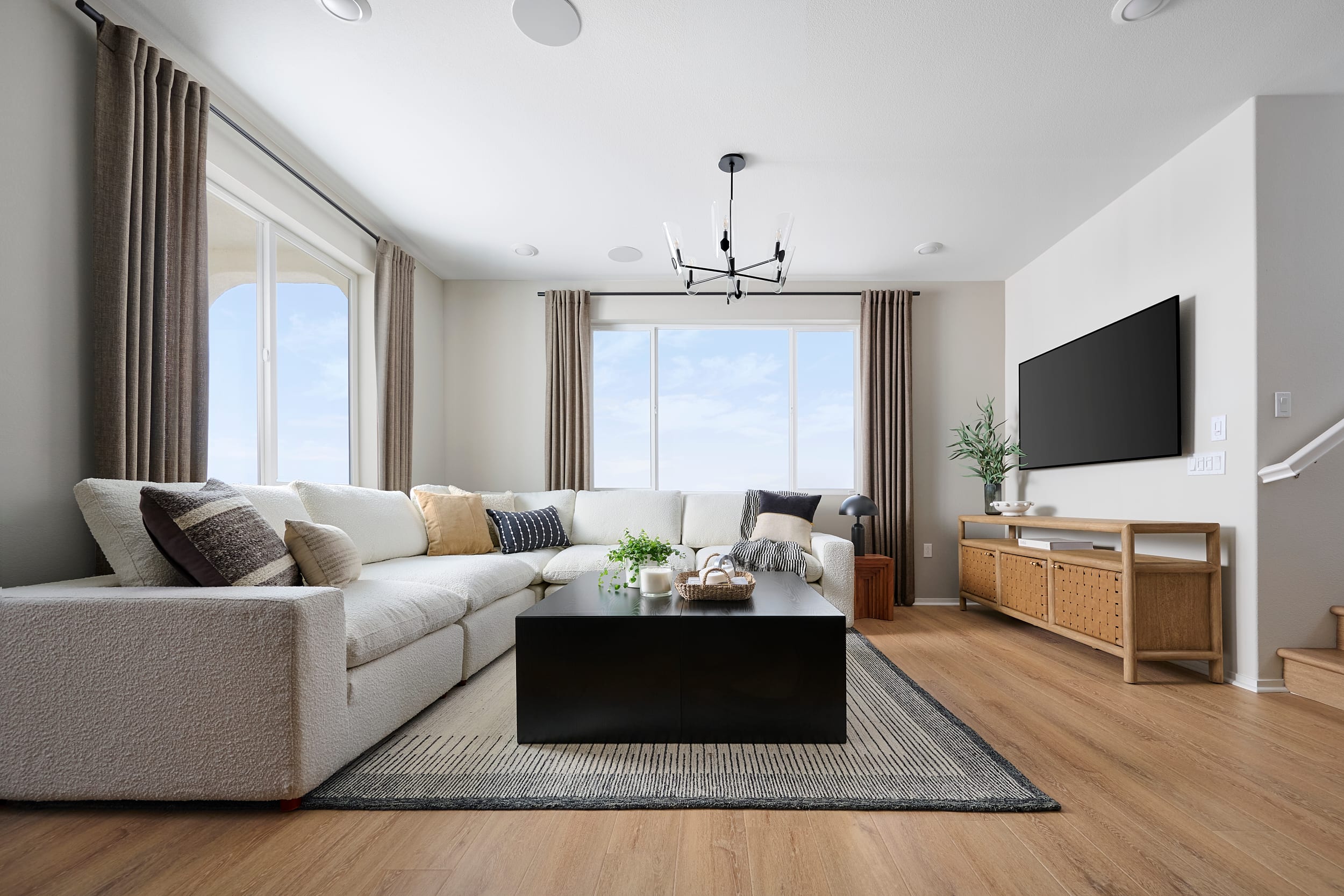 A large sectional sofa with decorative pillows sits in the foreground facing a black coffee table, while large windows with beige curtains and a television on a woven stand fill the background.