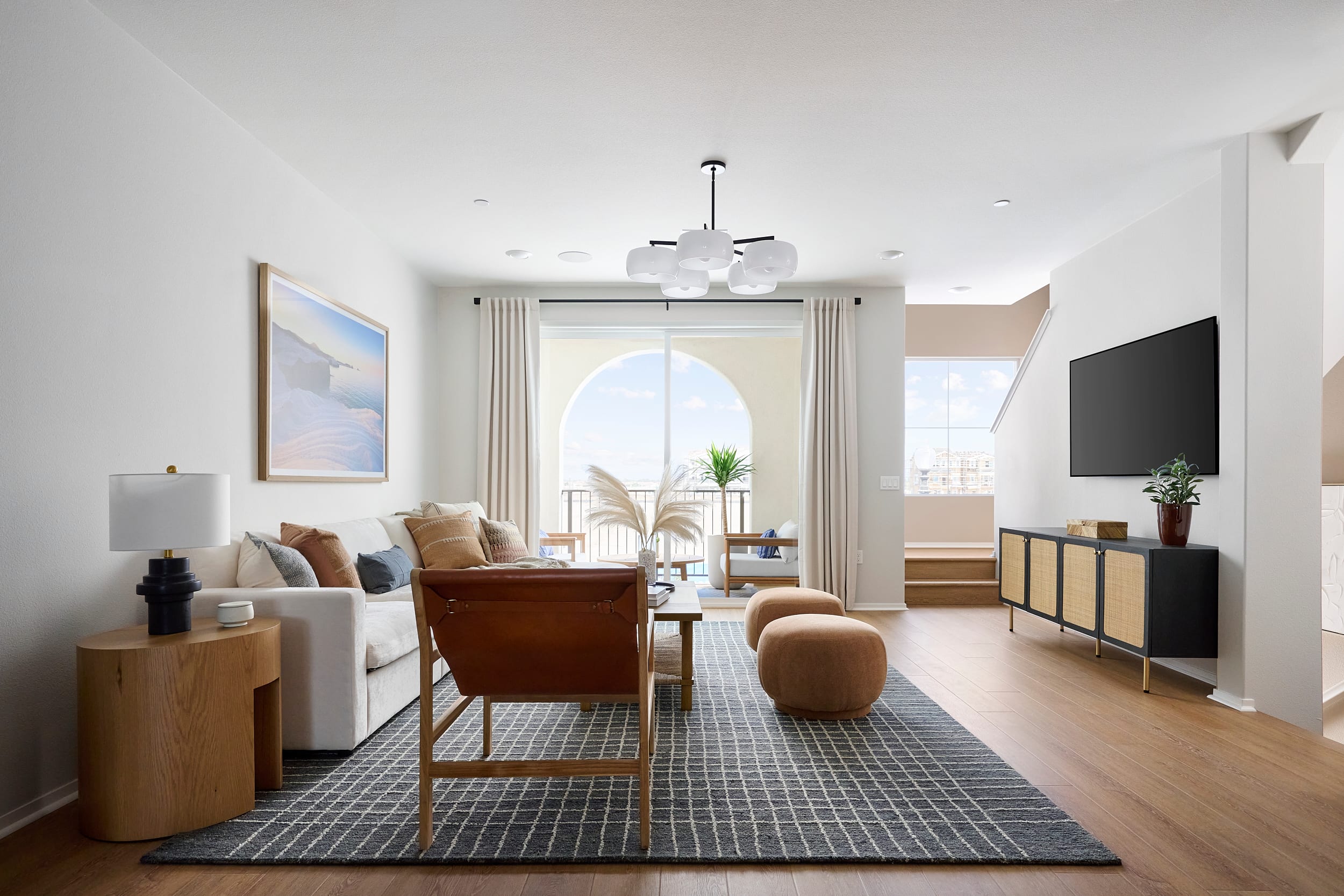 A modern living room with a white sofa and leather chair in the foreground, and a large arched window with a view of a balcony and distant buildings in the background.