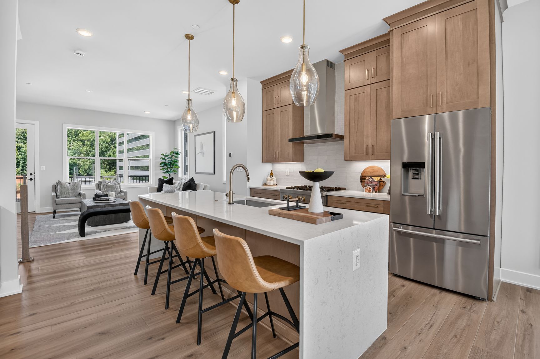 A modern kitchen island with bar stools in the foreground opens to a living area with large windows in the background.