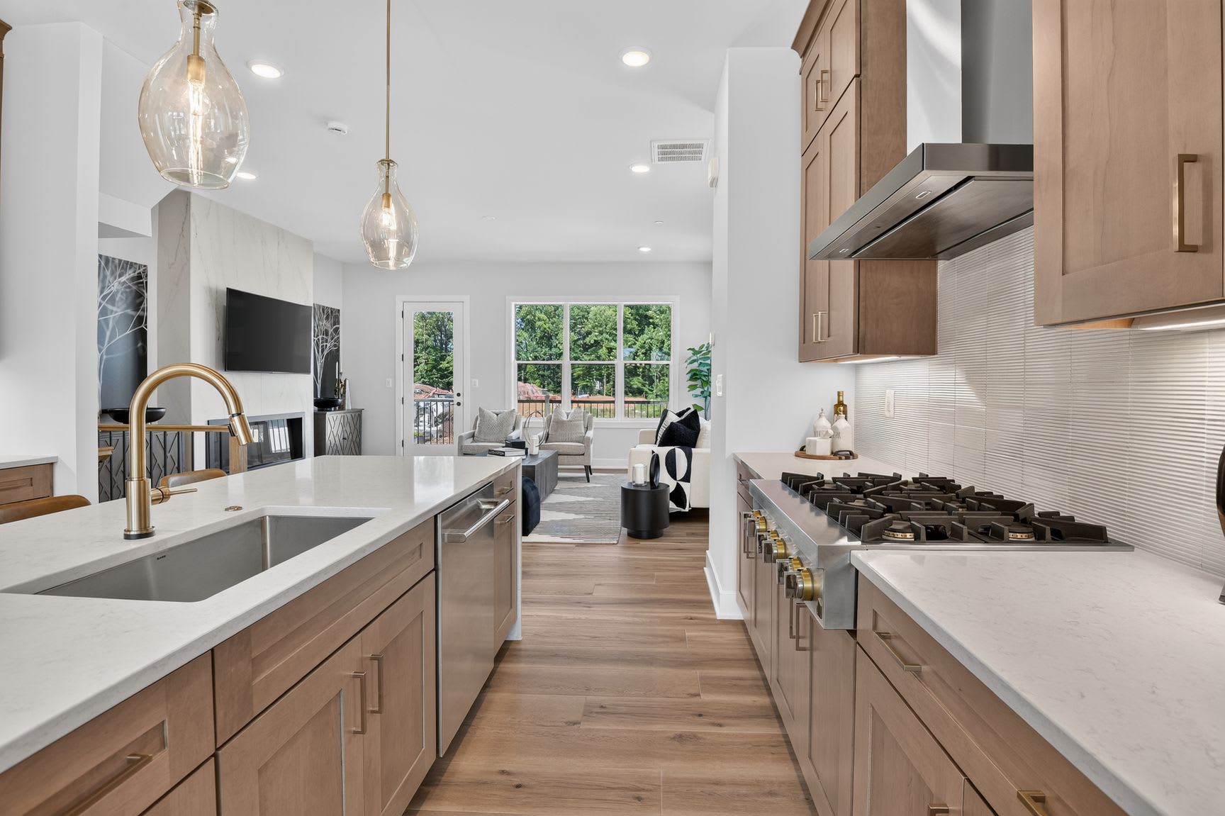 A modern kitchen with a large island and stainless steel appliances in the foreground opens to a living area with a fireplace and large windows in the background.