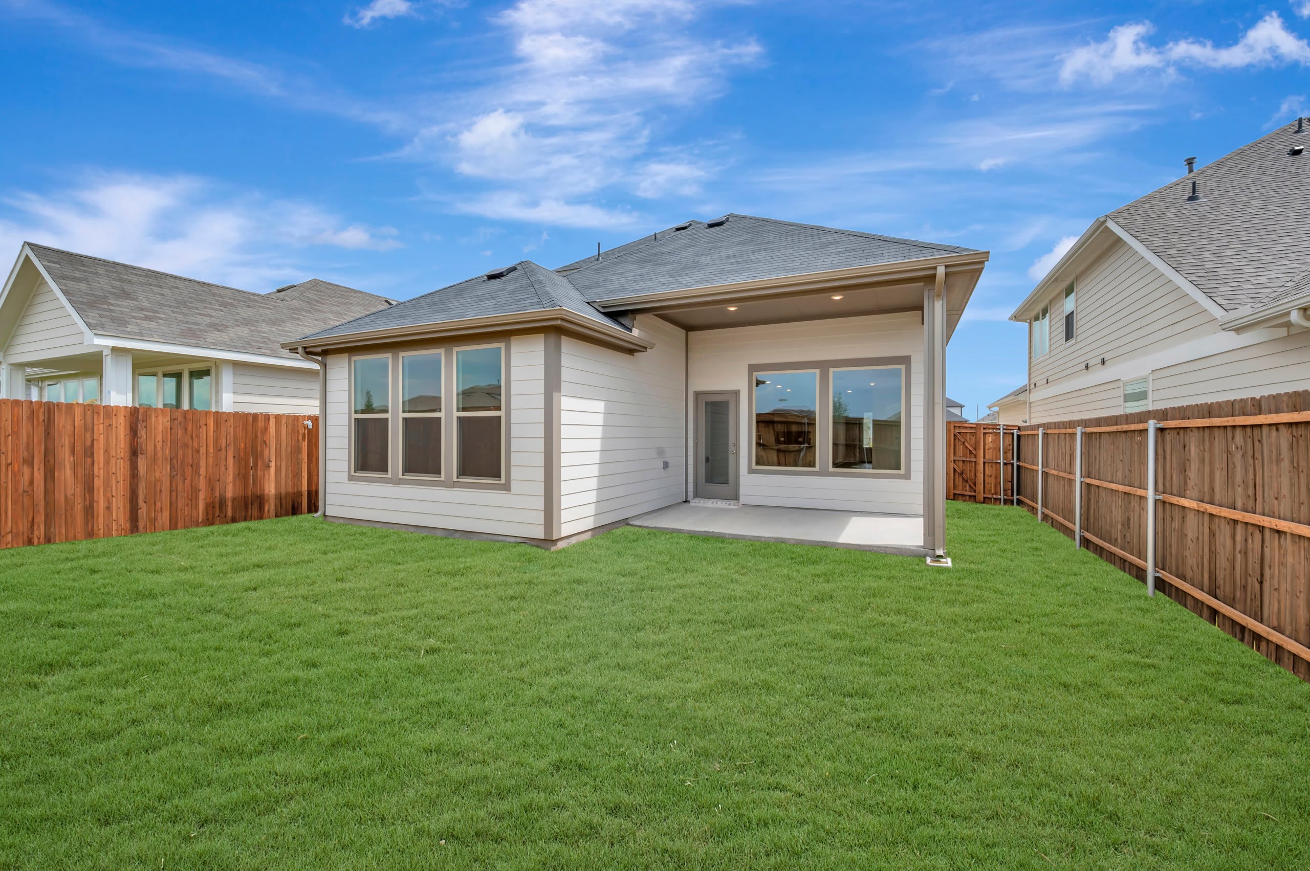 A freshly mowed green lawn fills the foreground, leading to the back of a modern house with large windows and a covered patio, all enclosed by wooden fences under a bright blue sky with scattered clouds.