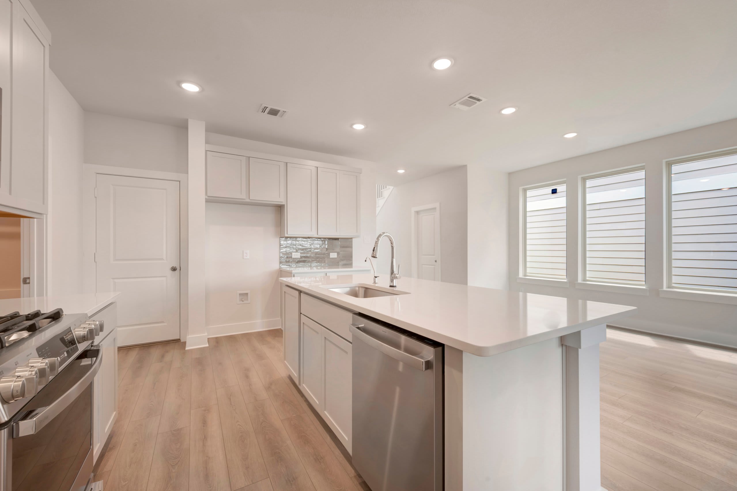 A modern white kitchen with a stainless steel stove in the foreground and a bright living area with large windows in the background.