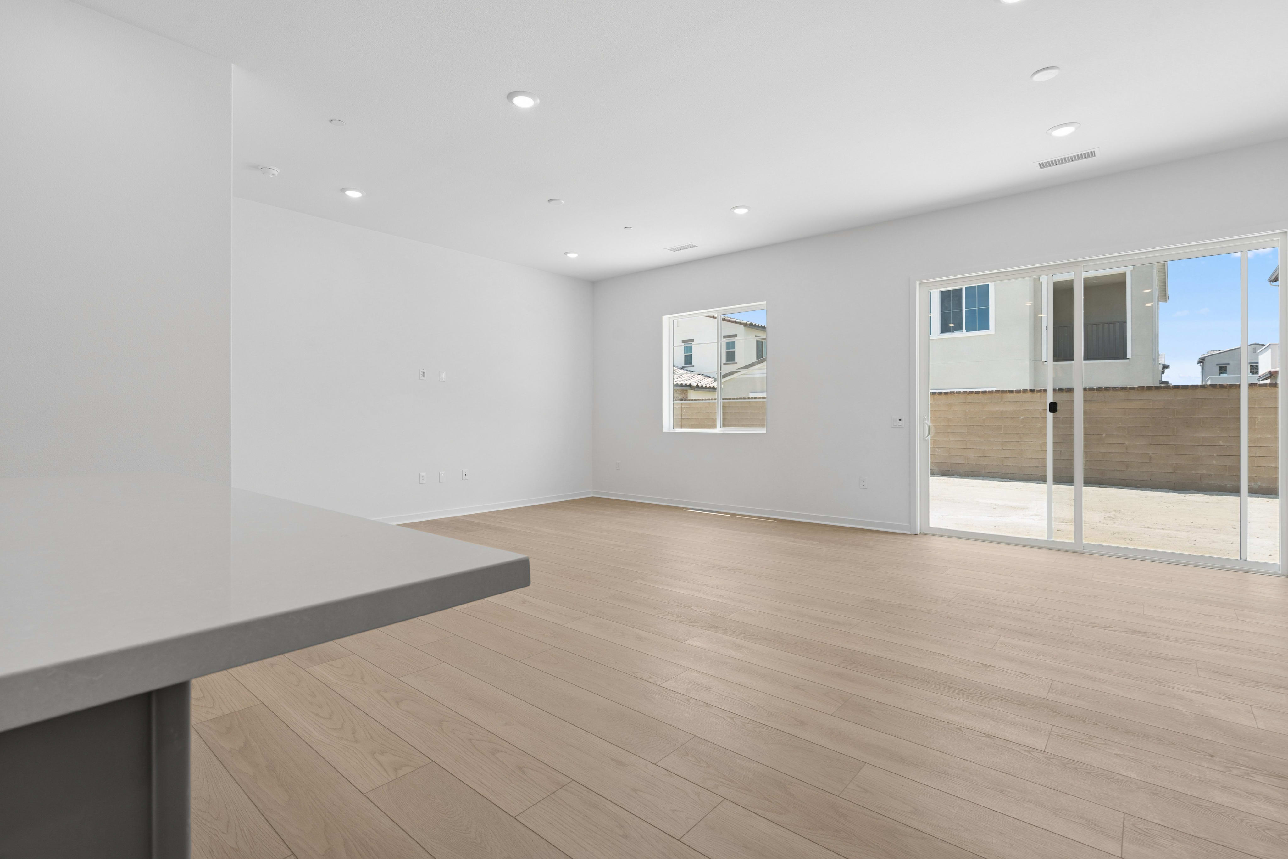 A modern living room with a light wood floor and white walls opens to a patio with a brick wall and houses visible in the background.