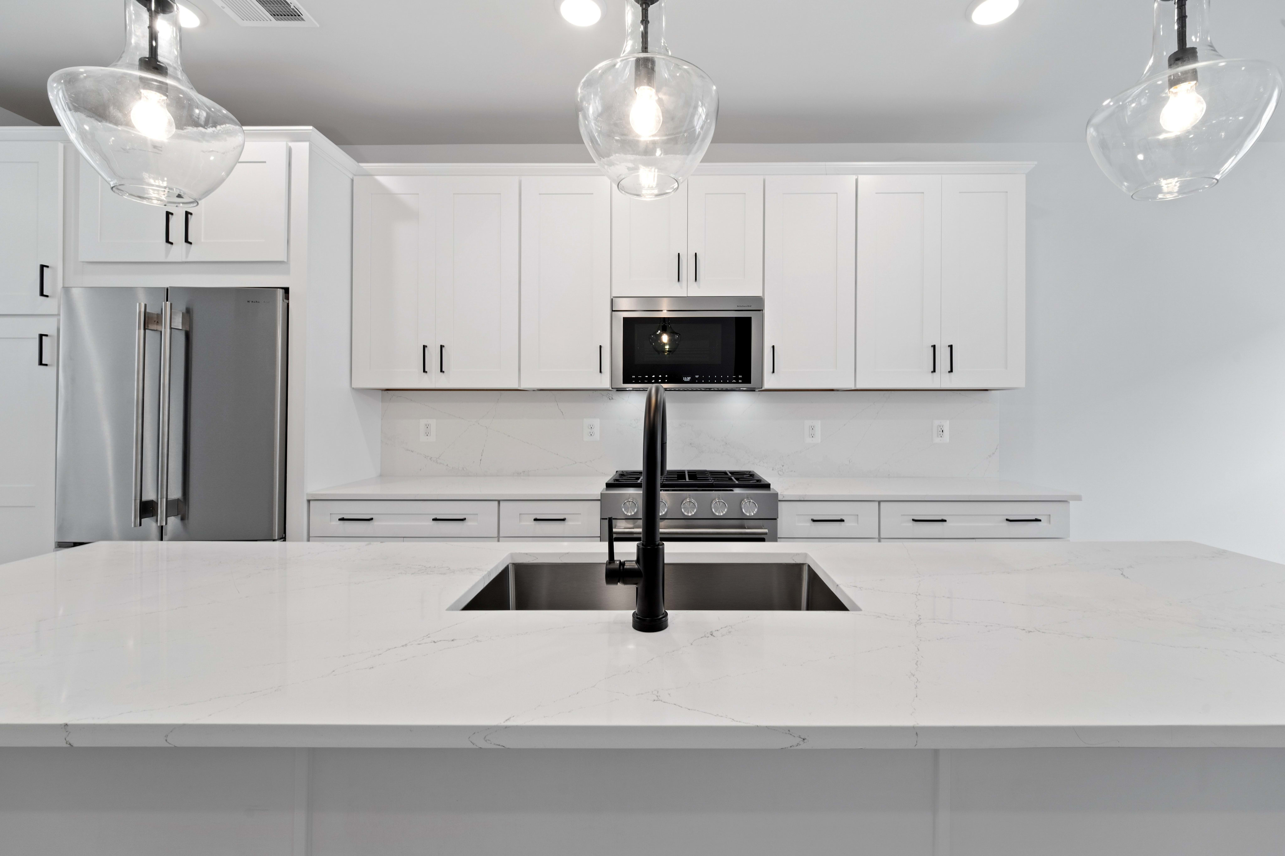 A modern white kitchen island with a black faucet and sink is in the foreground, while white cabinets, a stainless steel microwave, and a stove are in the background.