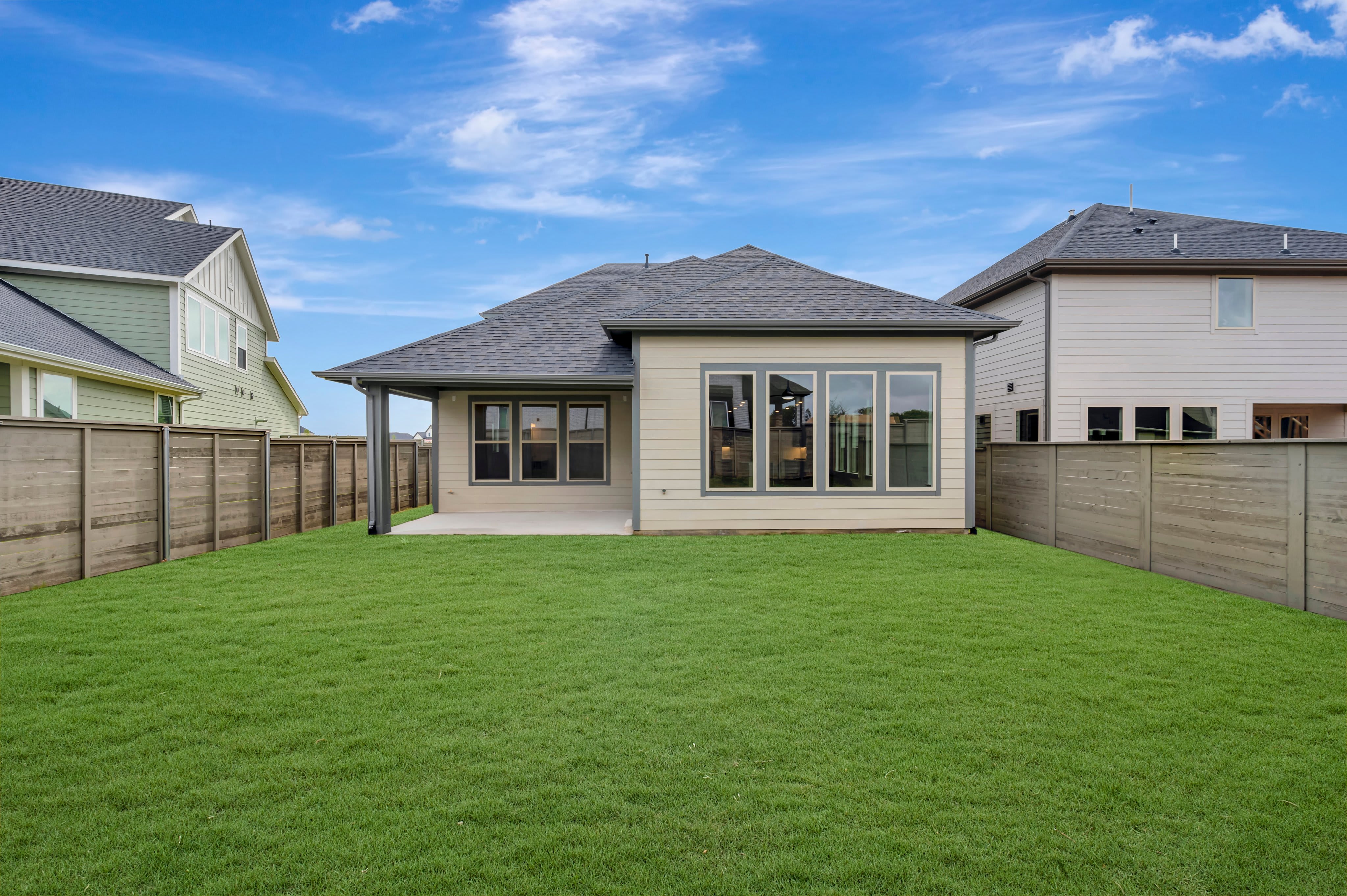 A lush green lawn fills the foreground, leading to the back of a beige house with large windows, while neighboring houses and a wooden fence form the background under a cloudy blue sky.