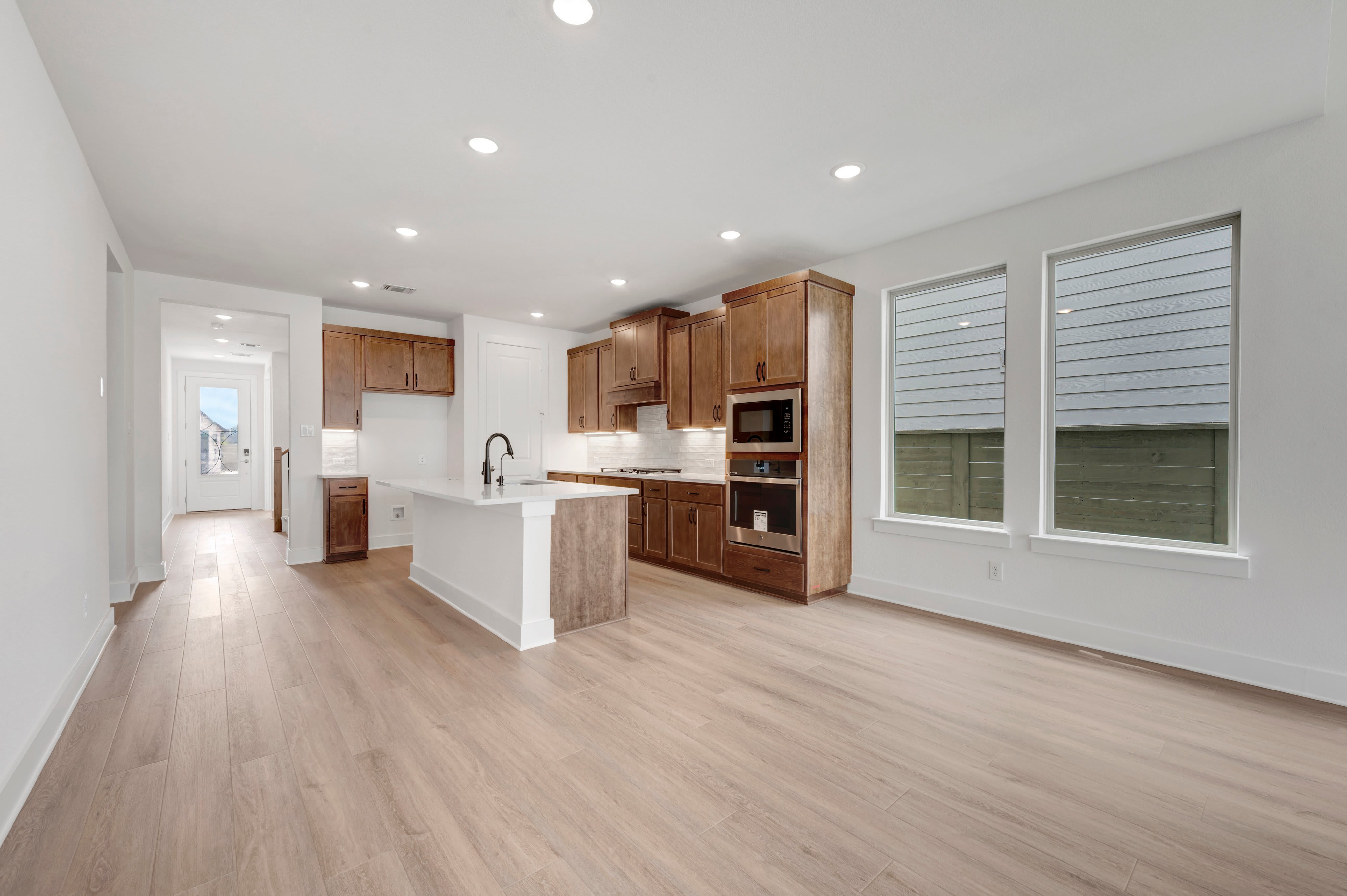 A modern kitchen with a white island and wooden cabinets is in the foreground, while a hallway and large windows overlooking a fence are in the background.