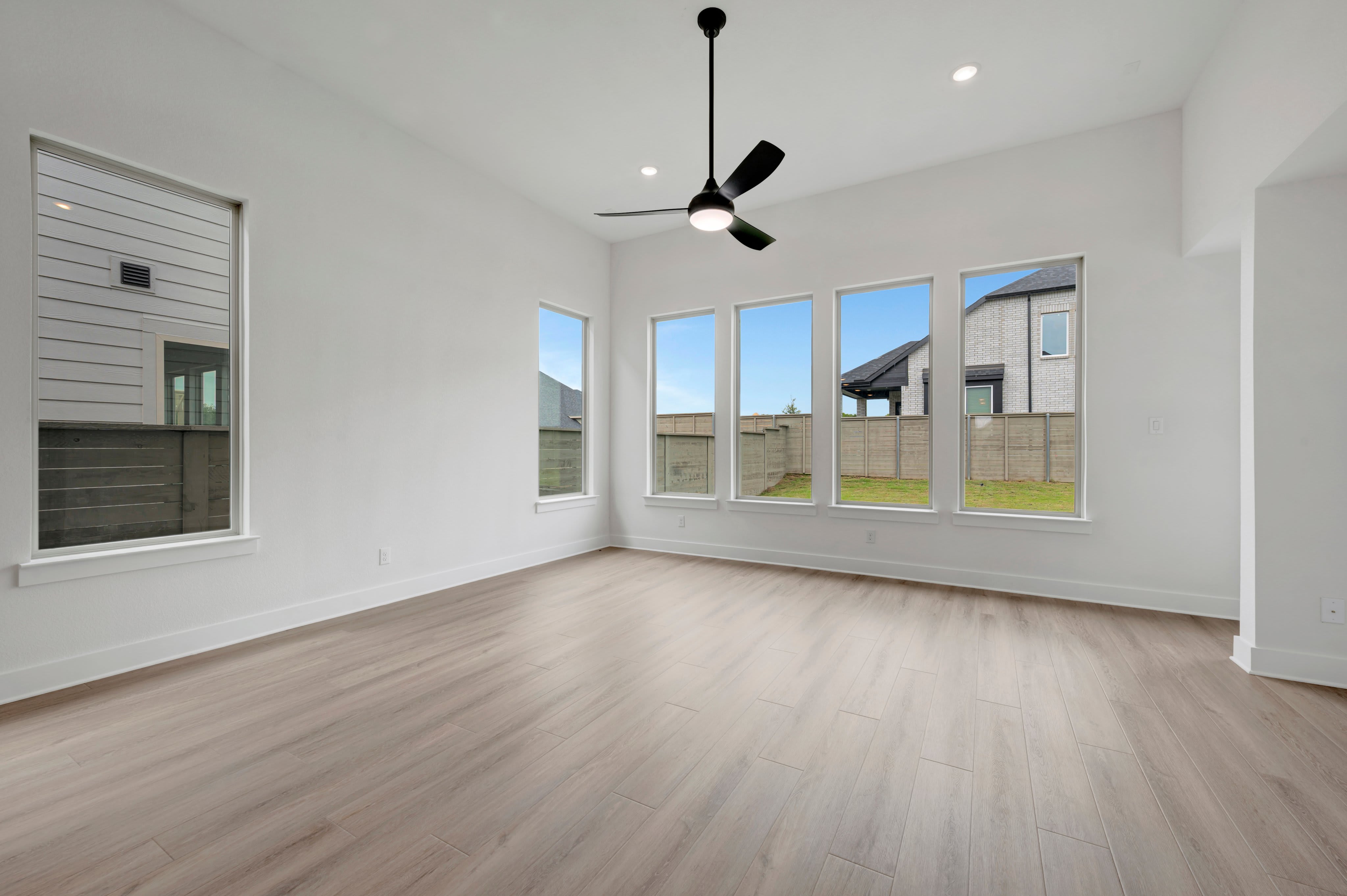 A spacious, modern living room with light wood floors in the foreground opens to a backyard with houses and a fence visible through large windows in the background.