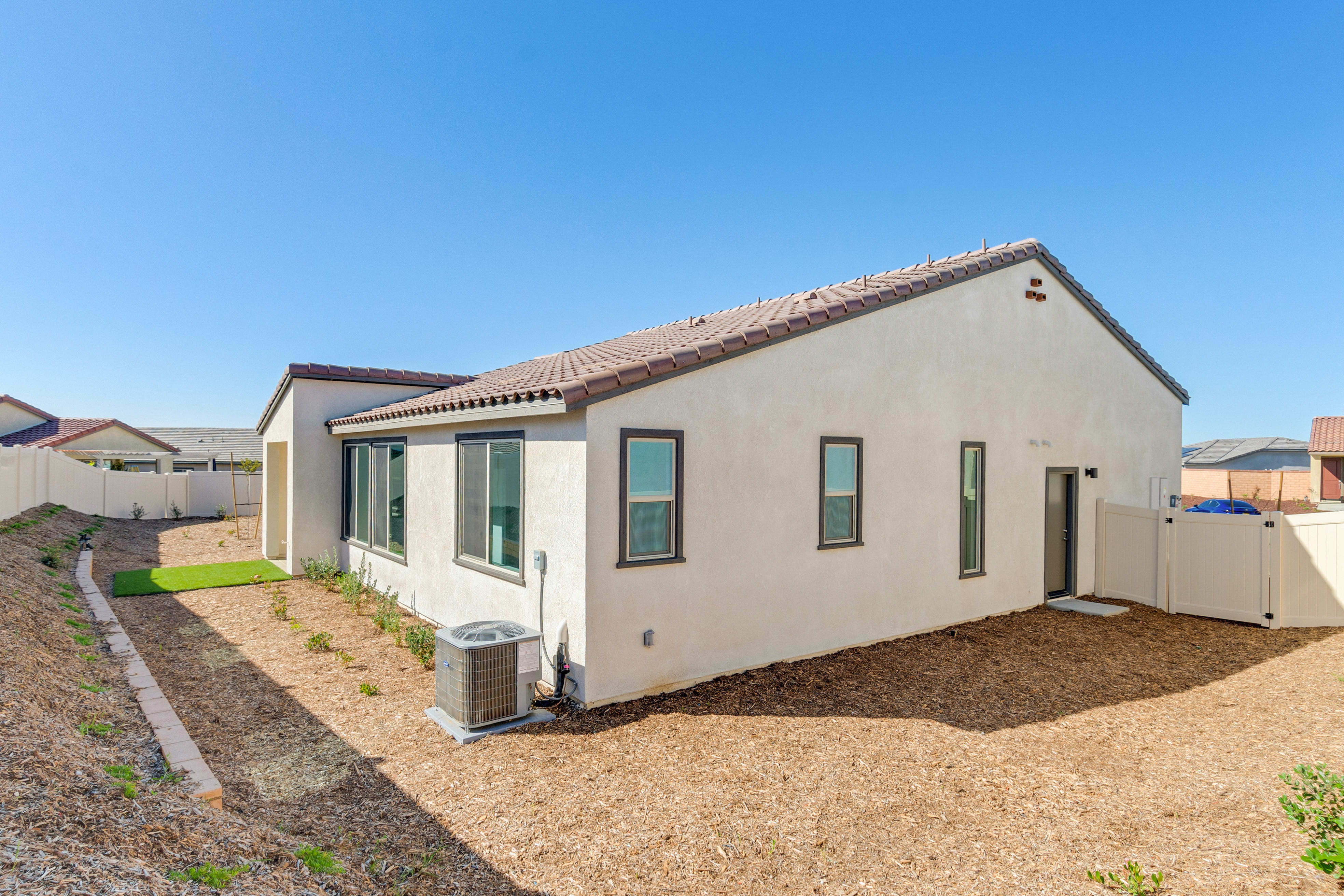 A white stucco house with a tile roof sits in a yard with wood chip mulch and a small patch of grass, backed by a clear blue sky and distant houses.