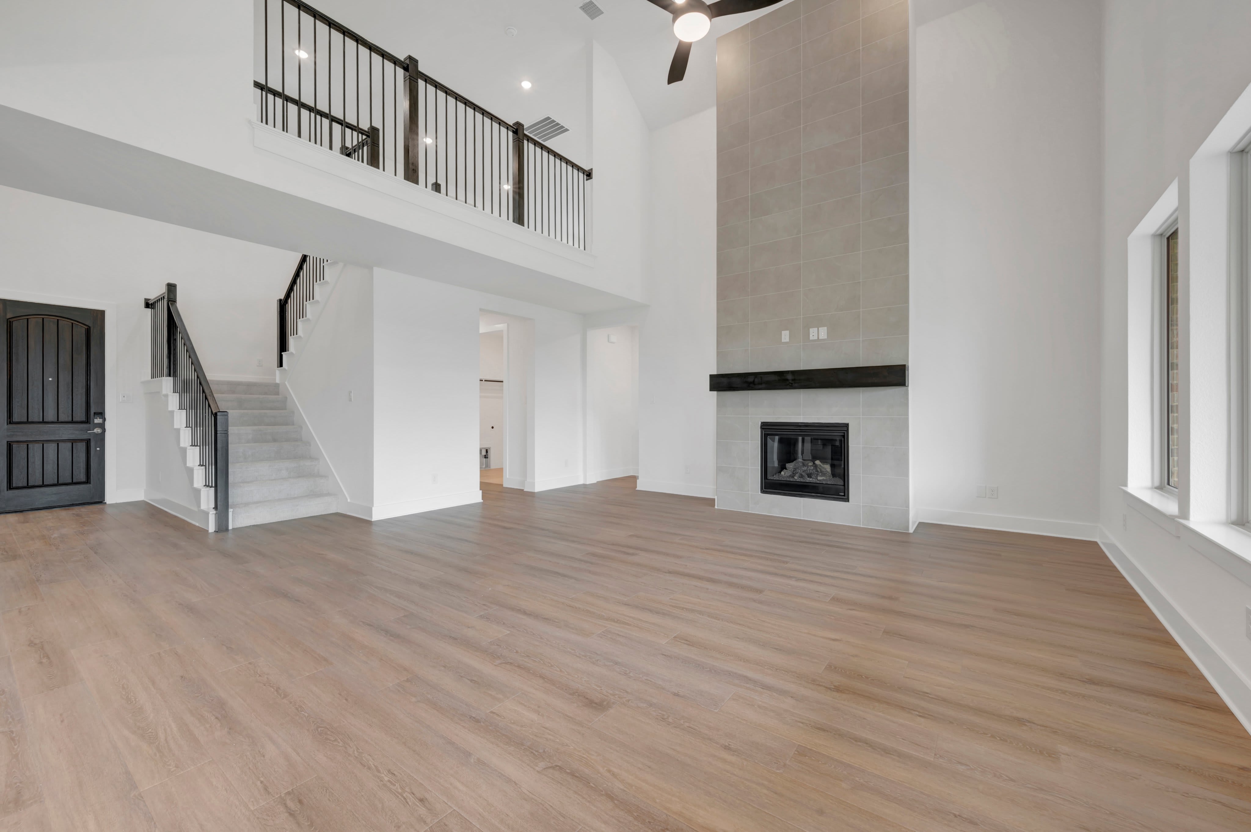 A spacious, modern living room with light wood floors in the foreground and a tall, gray tiled fireplace with a black mantel in the background.