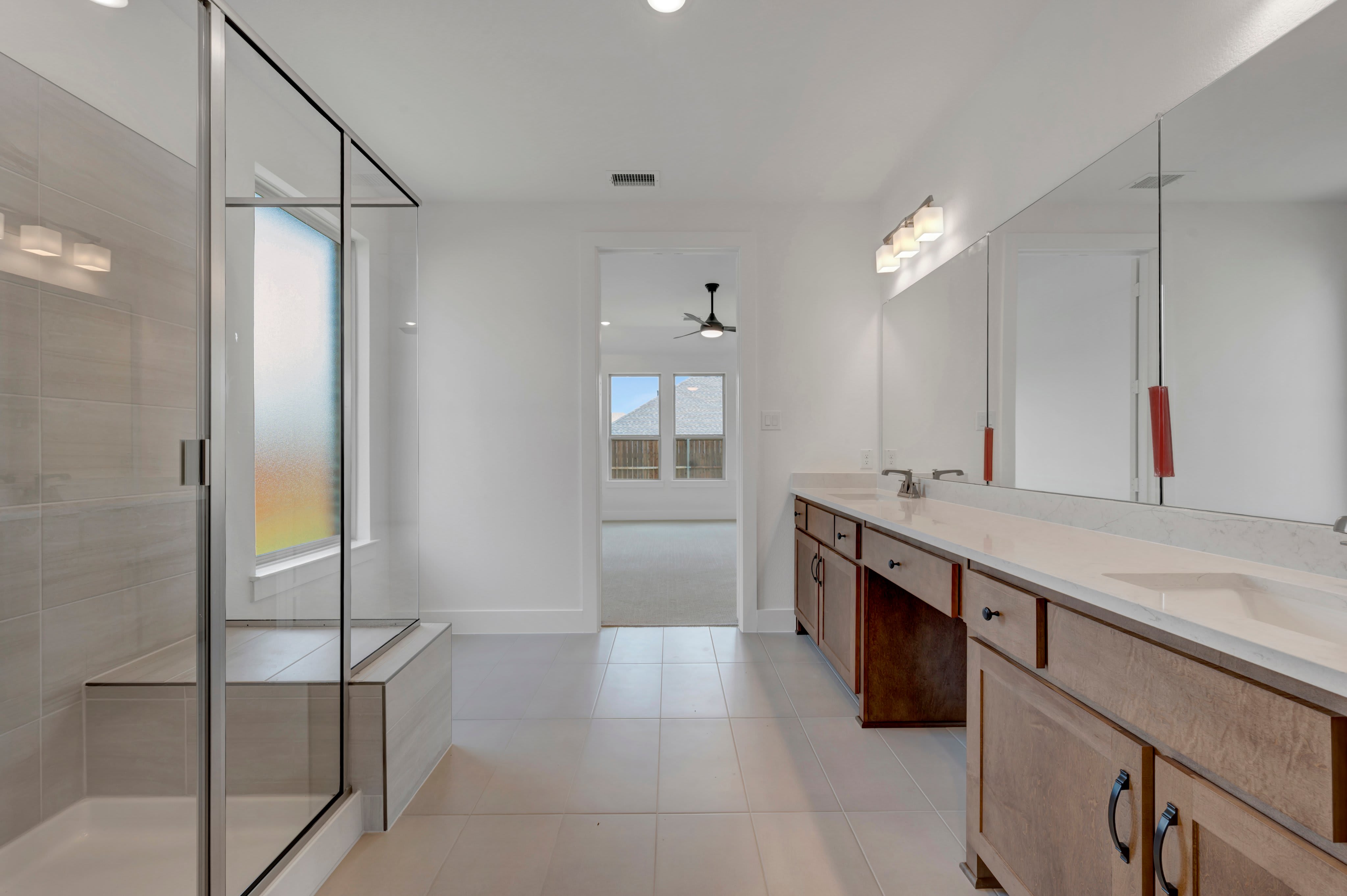 A modern bathroom with a glass-enclosed shower in the foreground and a long vanity with mirrors in the background, leading to a bedroom with a ceiling fan.