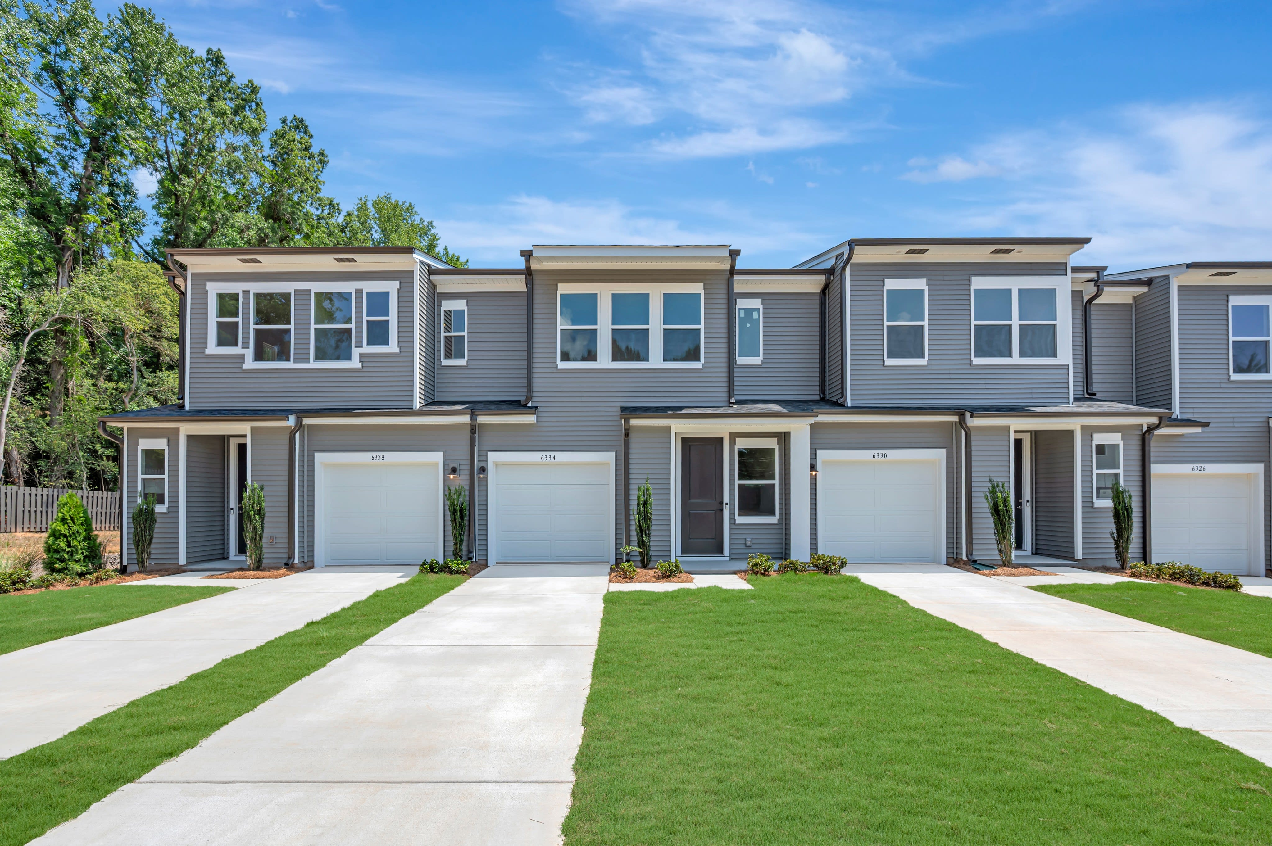 Modern gray townhouses with attached garages and manicured lawns sit beneath a bright blue sky with scattered clouds, backed by a line of lush green trees.