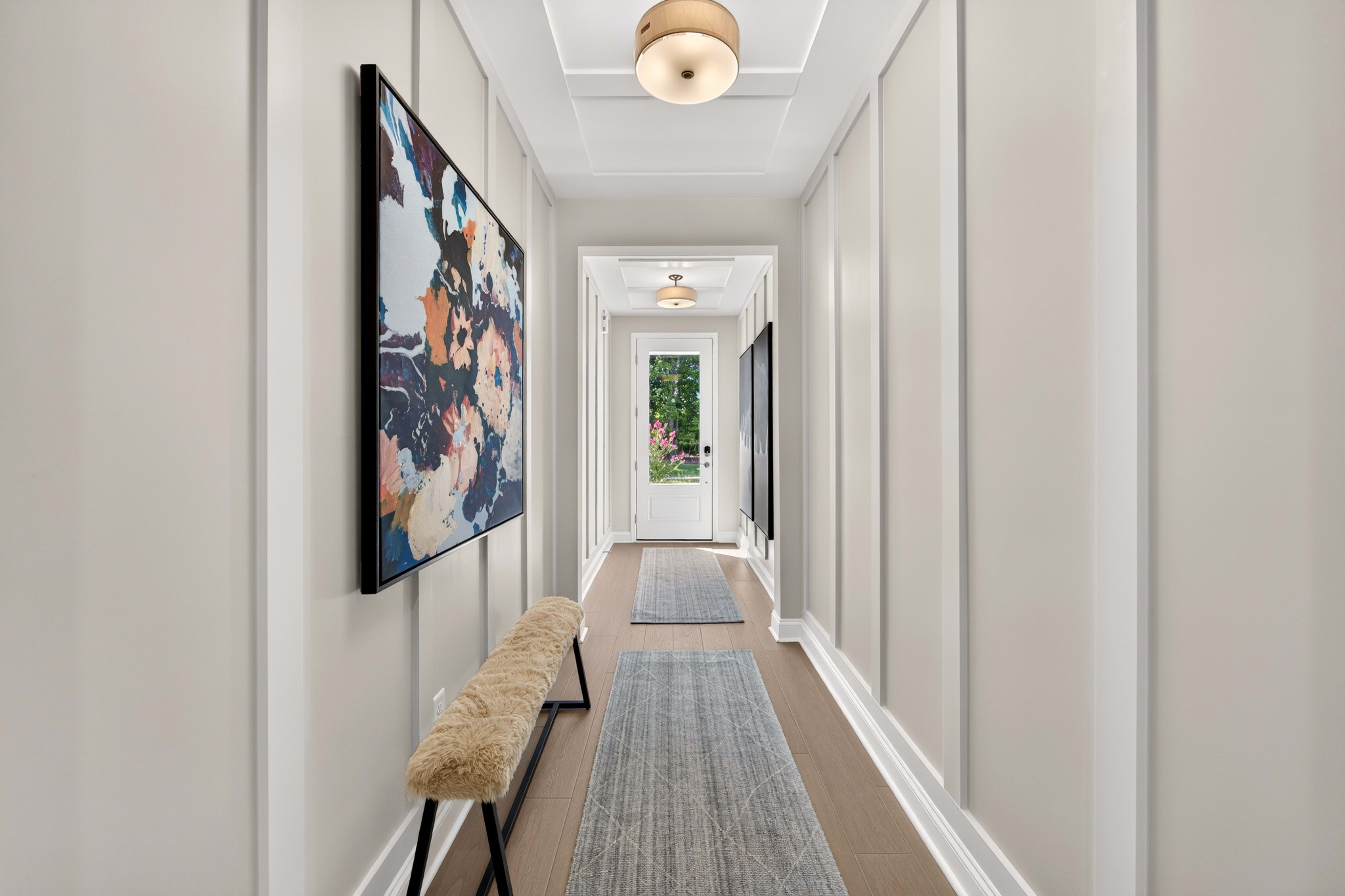 A modern hallway features a textured bench and abstract art in the foreground, leading to a bright doorway with greenery visible in the background.