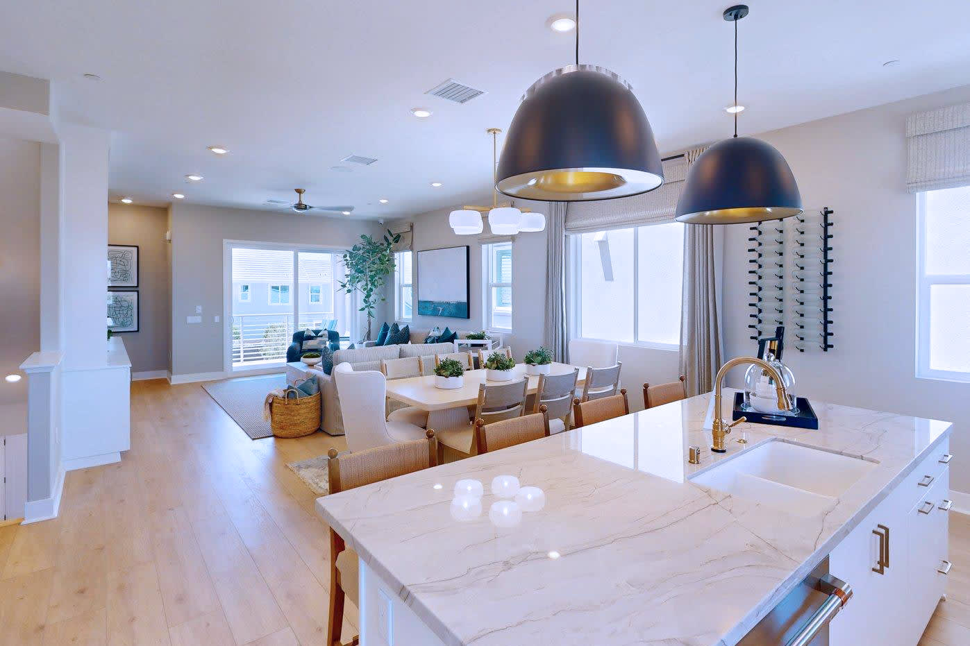 A modern kitchen island with a marble countertop is in the foreground, while a spacious dining and living area with large windows and contemporary furniture fills the background.
