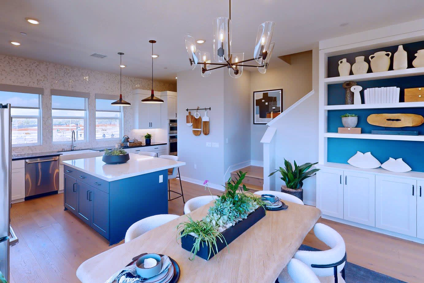 A modern kitchen and dining area with a blue island and a wooden table in the foreground, and a staircase and built-in shelves in the background.