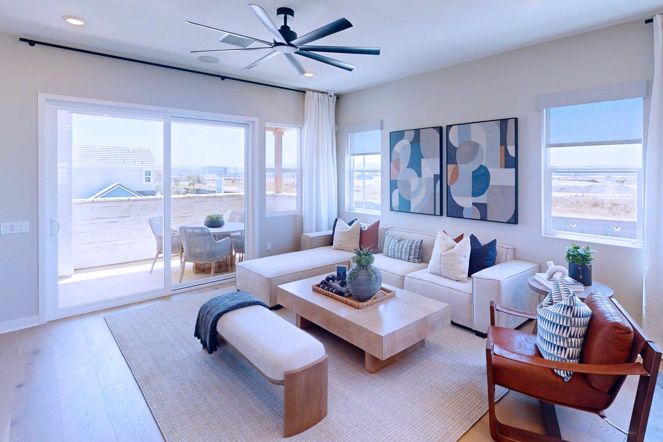 A modern living room with a sectional sofa and coffee table in the foreground, and a balcony with outdoor seating visible through sliding glass doors in the background.