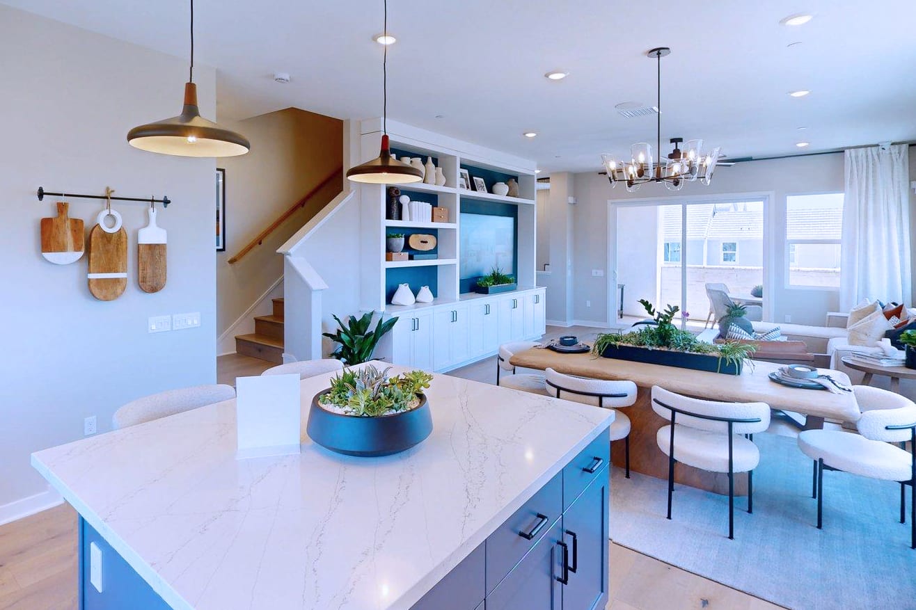 A modern kitchen island with a succulent arrangement in the foreground leads to a living area with a fireplace and large windows in the background.
