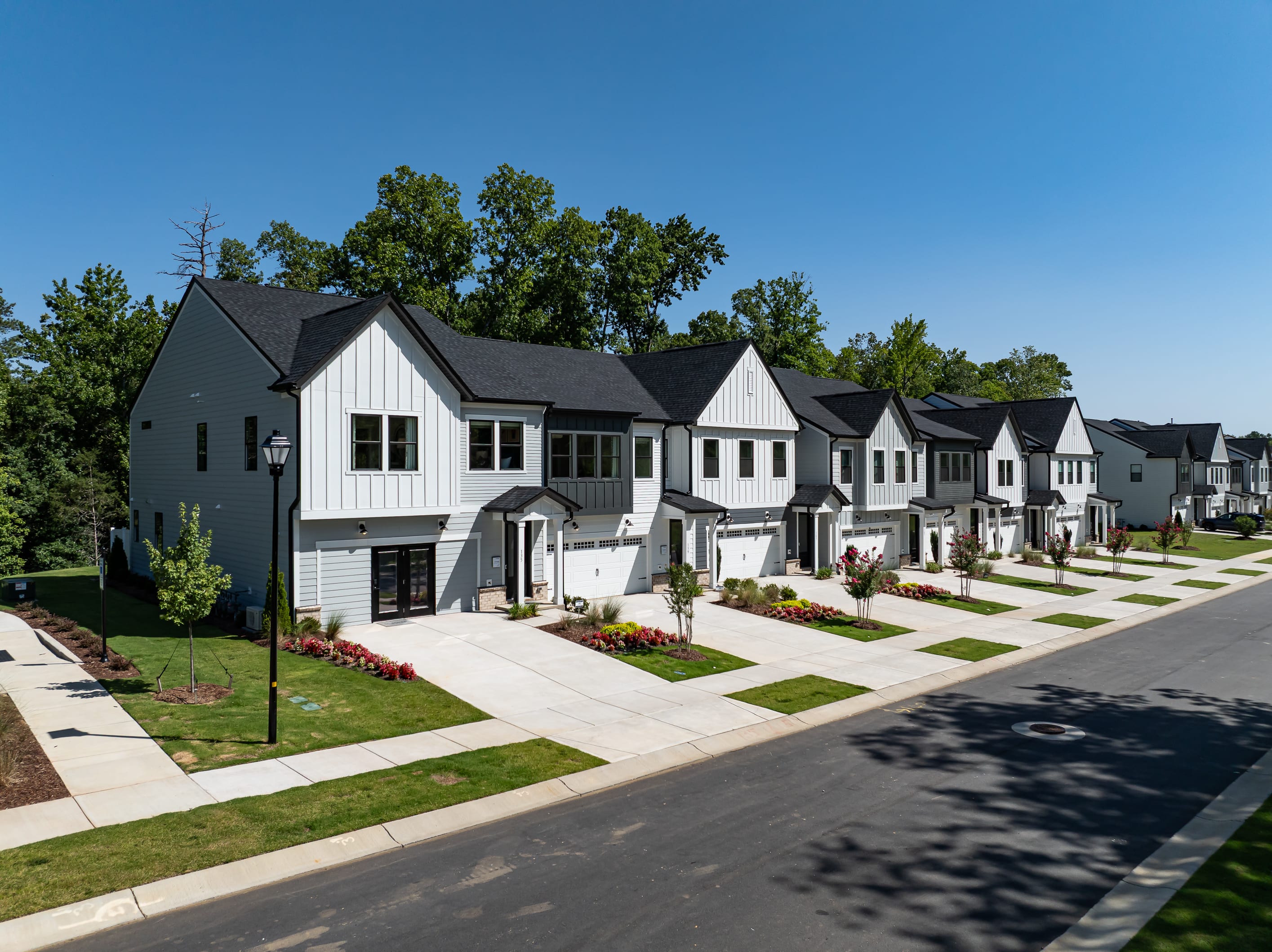 A row of modern townhouses with manicured lawns and driveways stretches towards a backdrop of lush green trees under a clear blue sky.